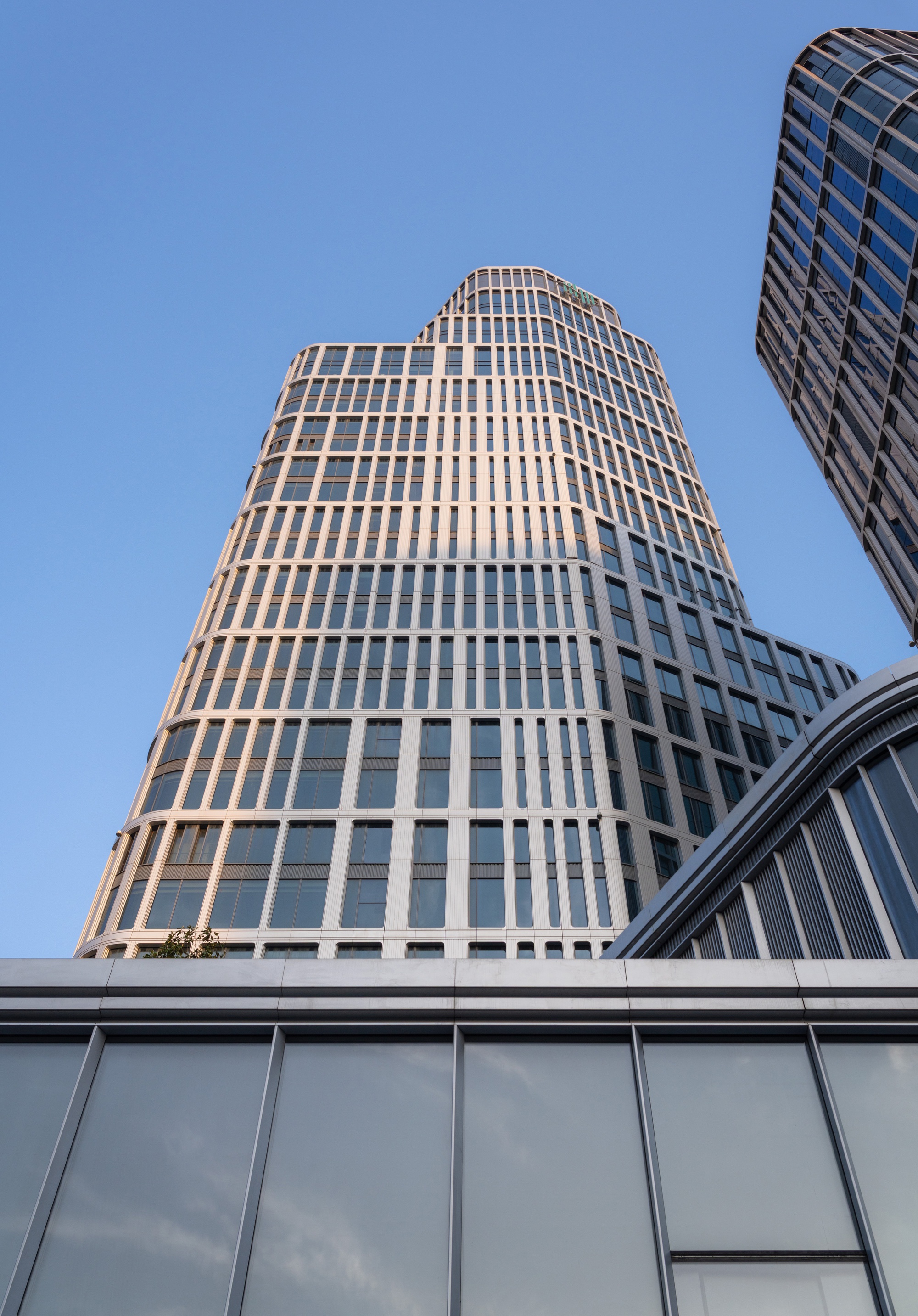 Low-angle view of a tapering tower with pale gridded facade and rounded corners against a clear blue sky