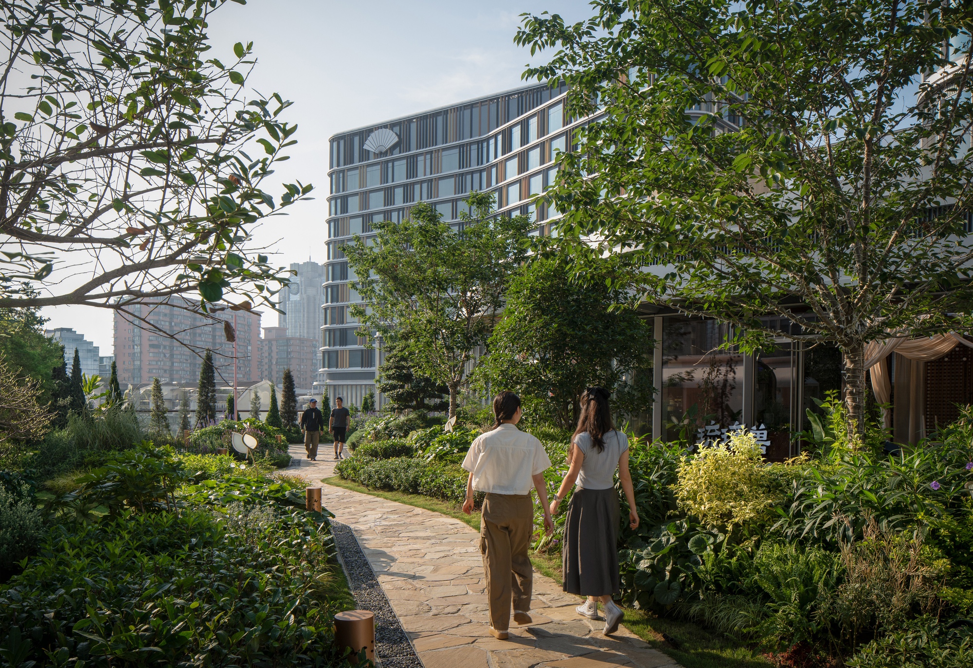 Two visitors walk a flagstone path through a rooftop garden of shrubs and trees toward a banded glass building