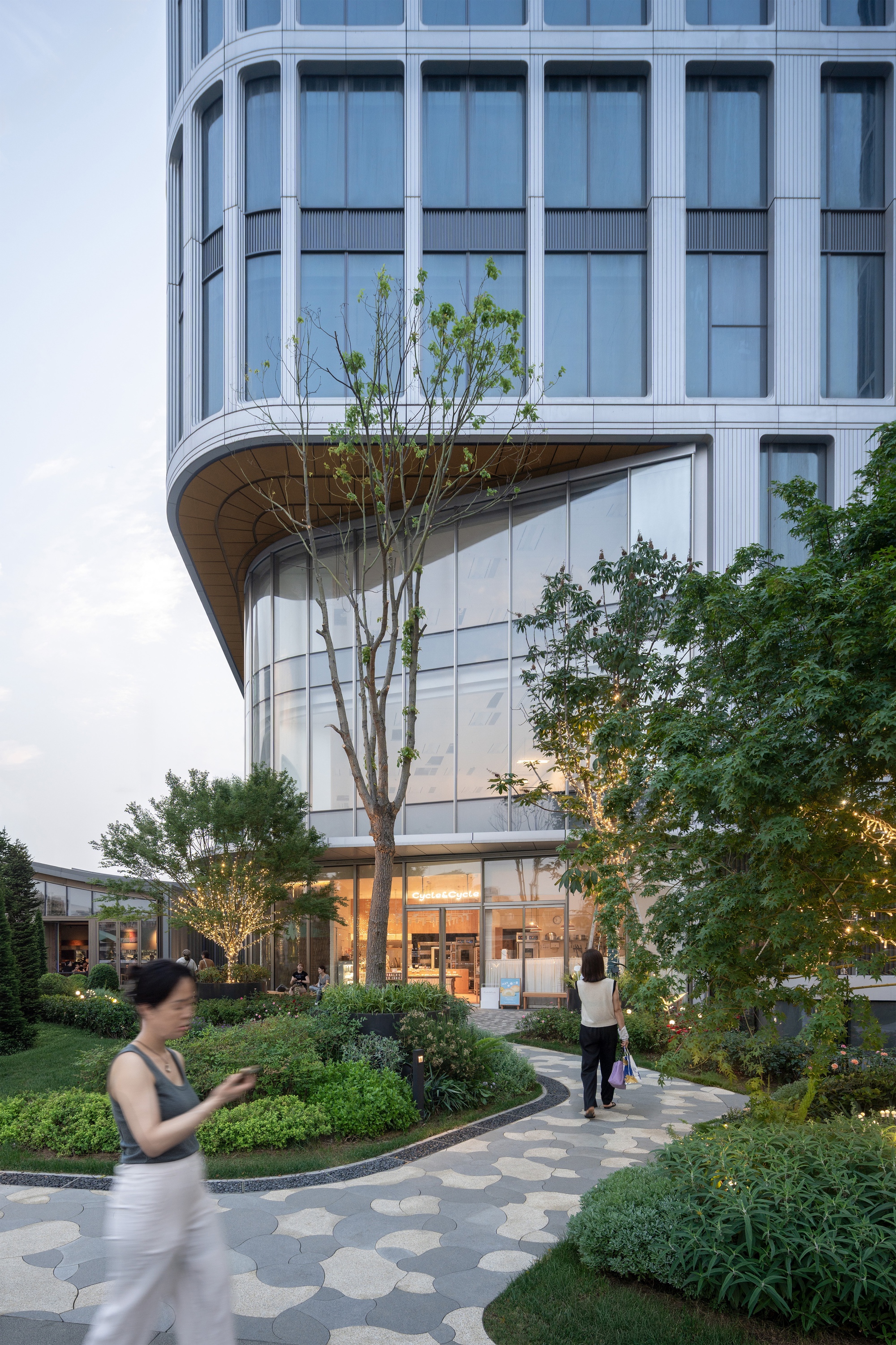 Curved timber-soffit corner of a tower rising above a landscaped courtyard with patterned paving and a woman walking past