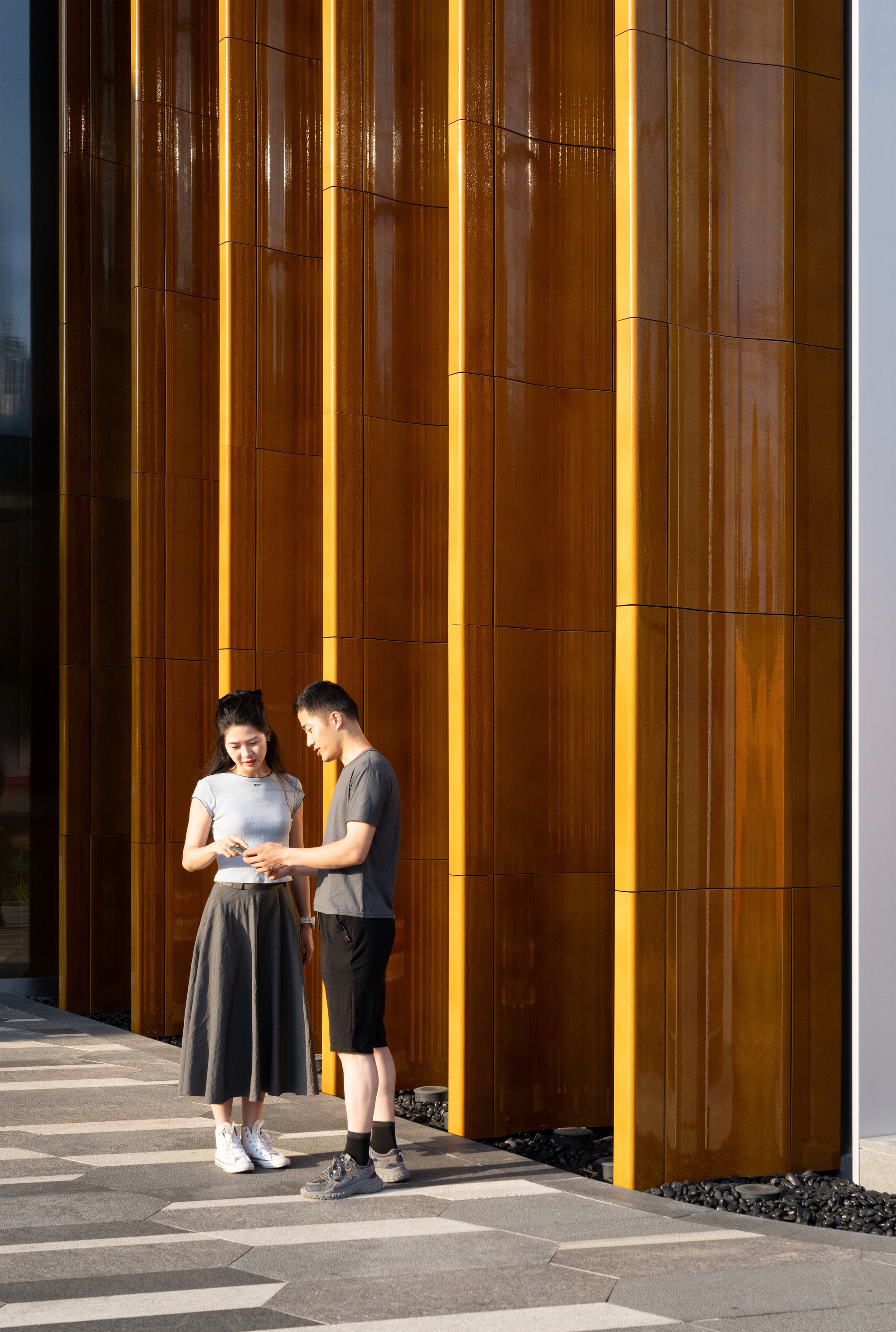 Two people stand reading a phone against a wall of vertical rust-colored metal fins in low golden sunlight