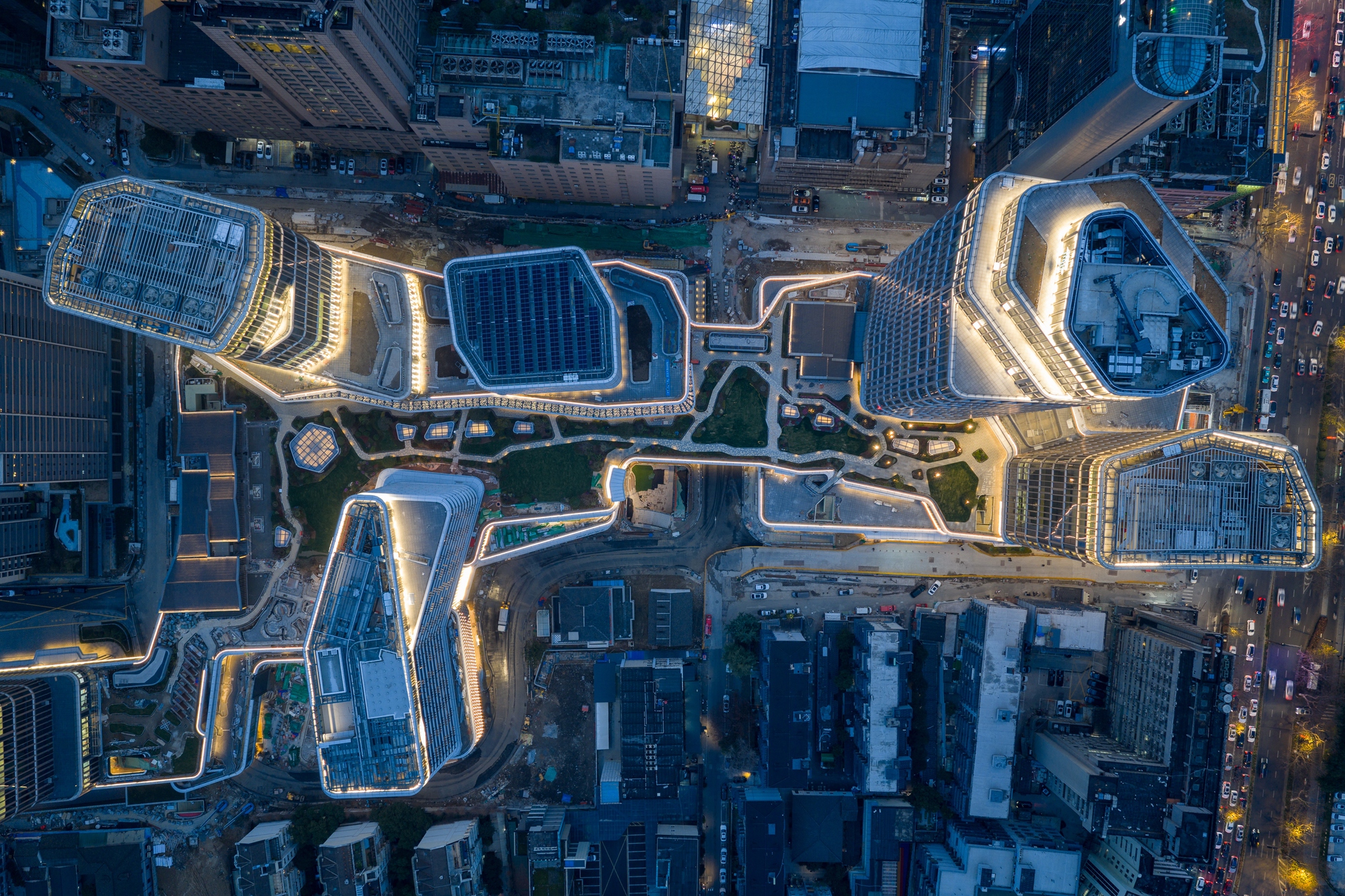 Aerial view of towers and linked rooftop terraces outlined in warm light over surrounding city blocks and traffic at dusk