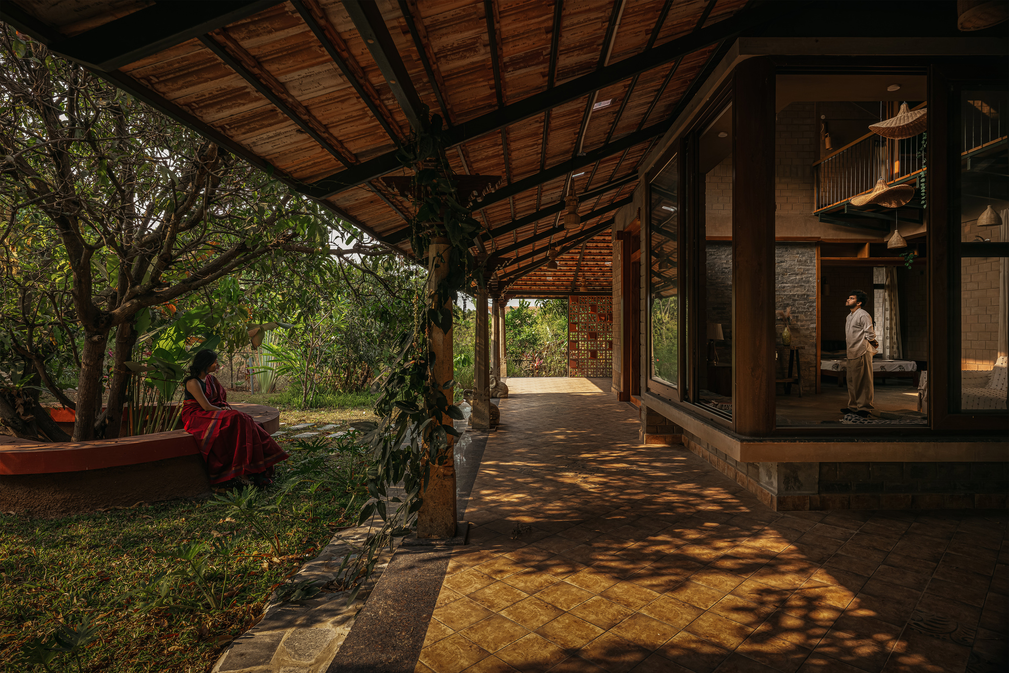 Covered veranda with timber posts and tiled roof, dappled sunlight on the floor and a woman seated on a curved bench