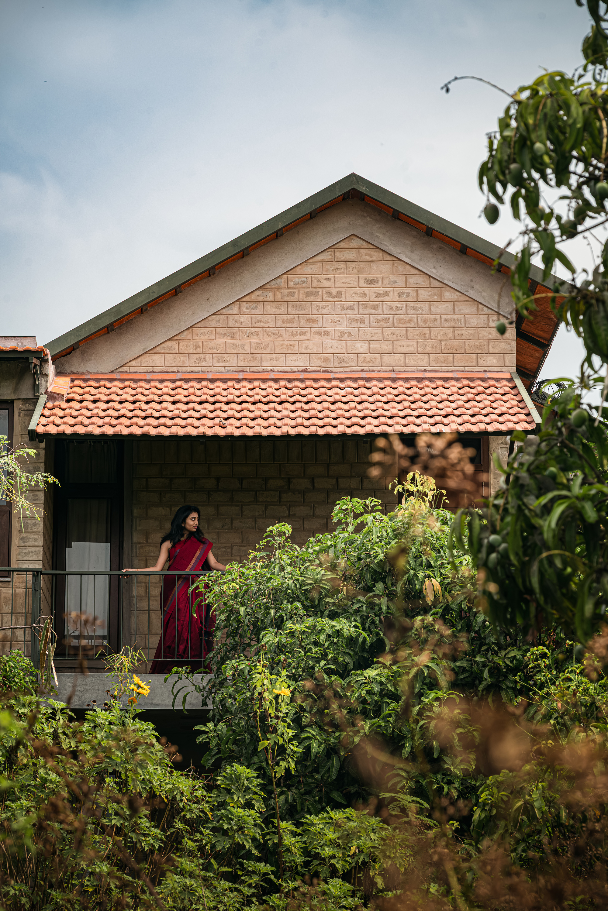Brick gable end with a tiled roof and a balcony where a woman in a red sari stands behind leafy branches