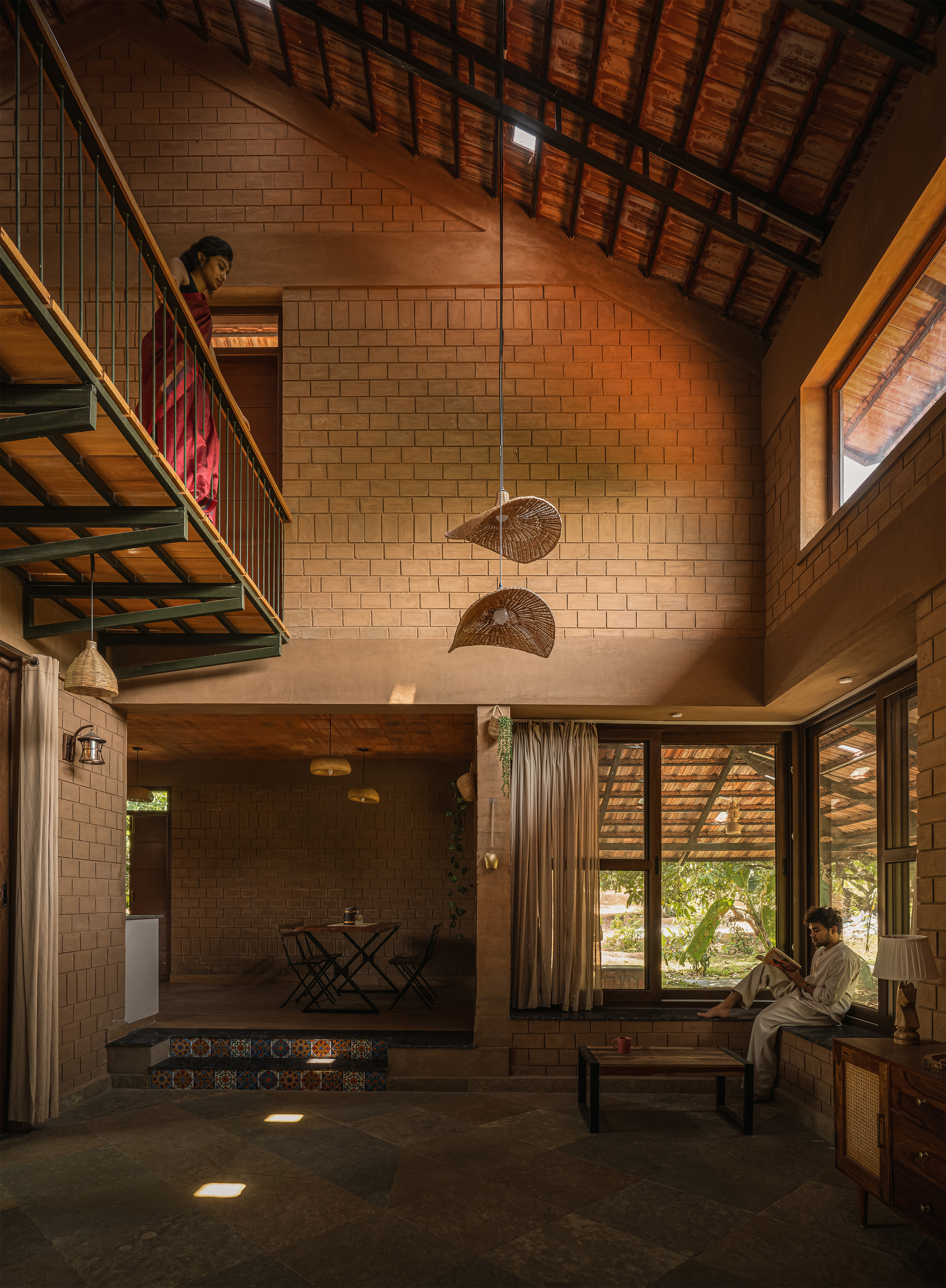 Double-height living space with brick walls, a steel and timber balcony, woven pendant lamps and a man reading by the window