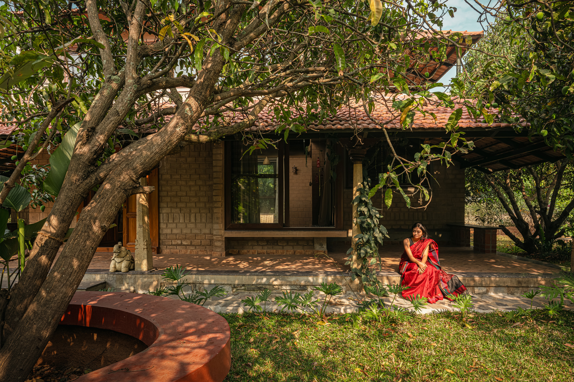 Brick house with a tiled roof and timber veranda seen through a tree trunk, a woman seated on the step in afternoon sun