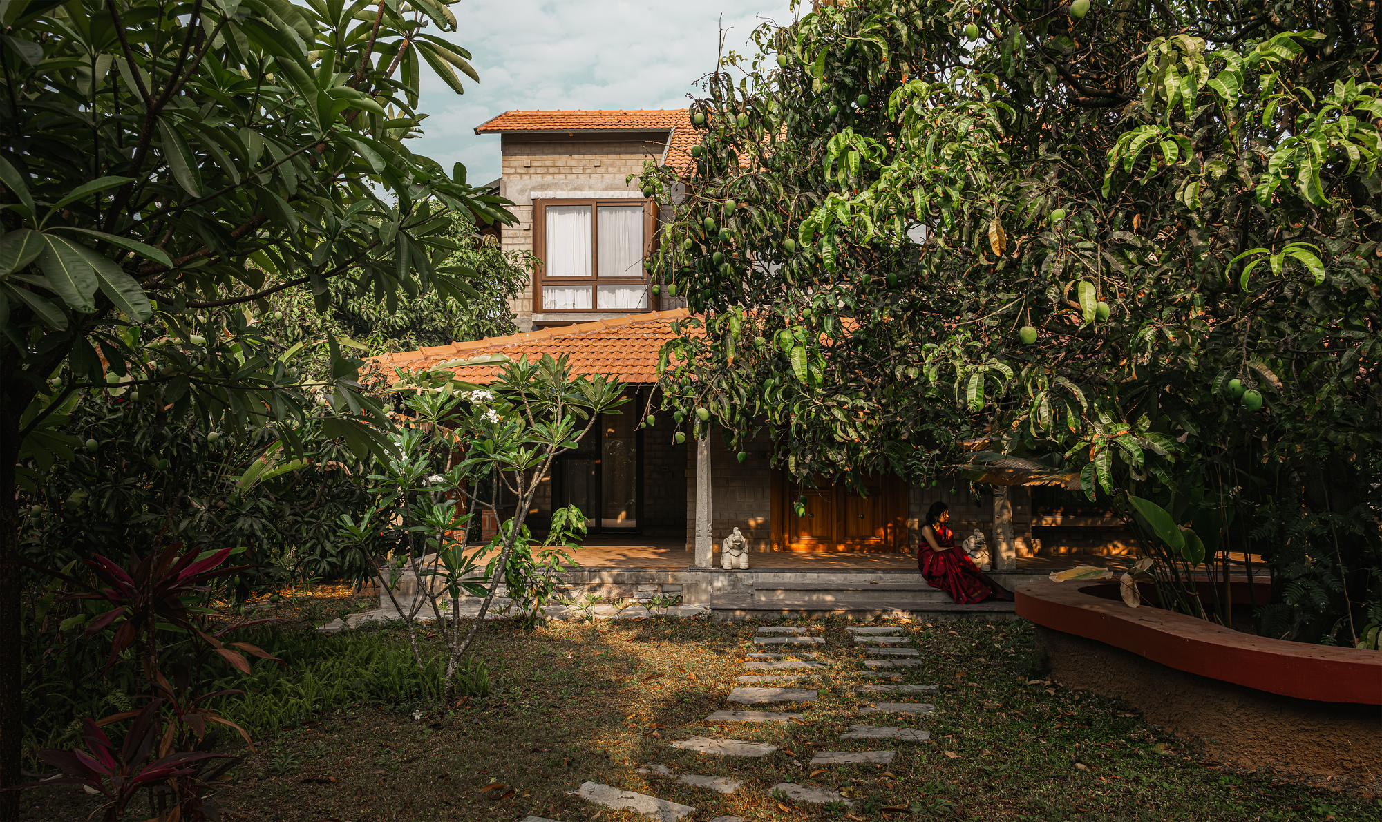 Brick house with terracotta tile roofs behind a stepping-stone path, framed by mango and frangipani trees in warm afternoon light