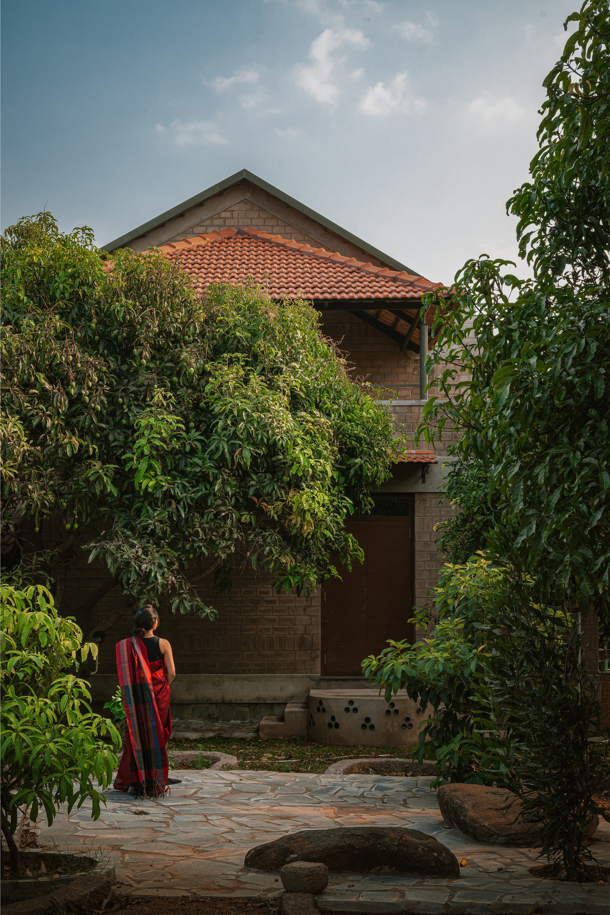 Brick facade with a pitched clay-tile roof and a perforated stone seat, seen across a flagstone courtyard with a woman in a red sari