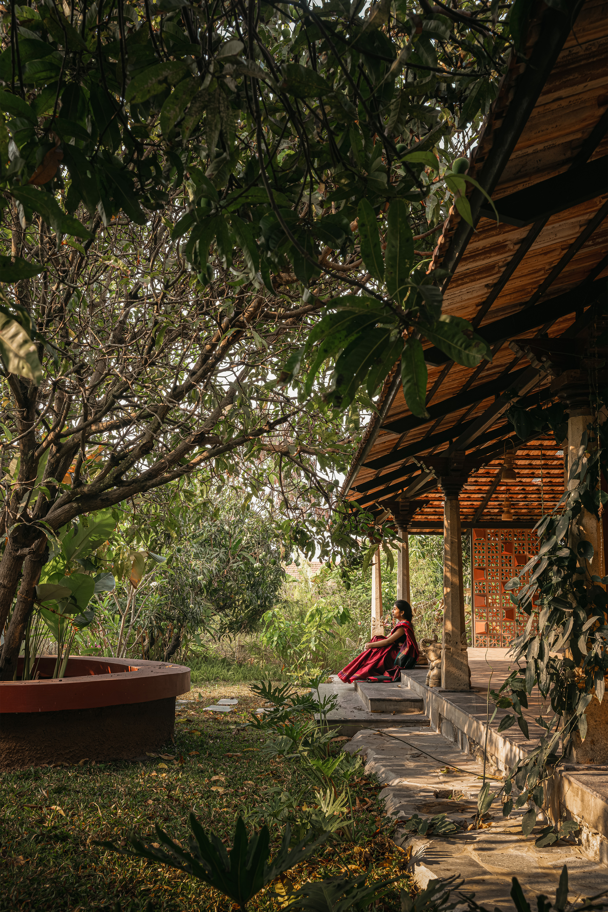 Timber-ceilinged veranda with carved columns and stone steps, a seated woman in red beneath a spreading tree canopy