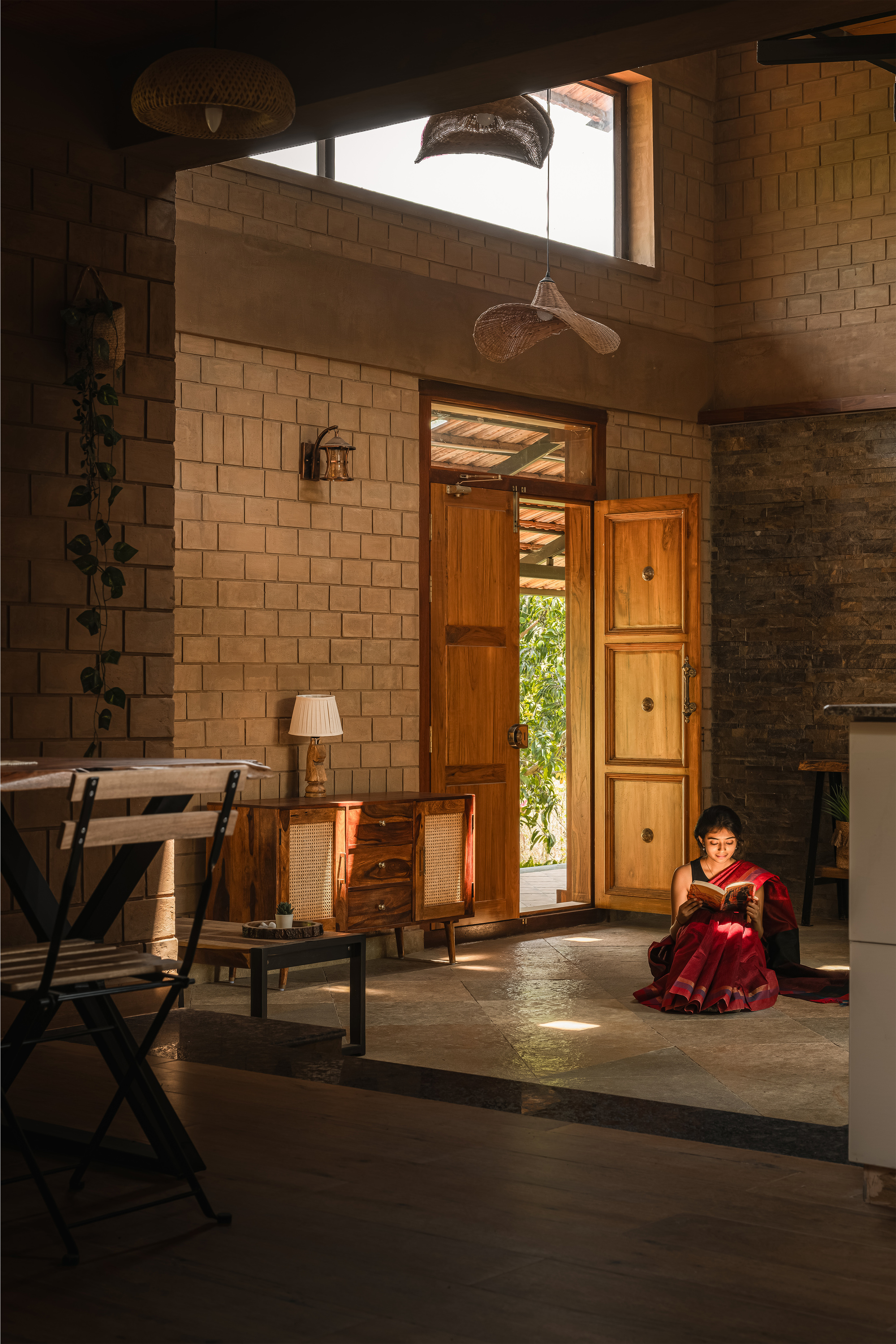 Living room with exposed brick walls and open teak doors, a woman reading on the stone floor in a shaft of sunlight