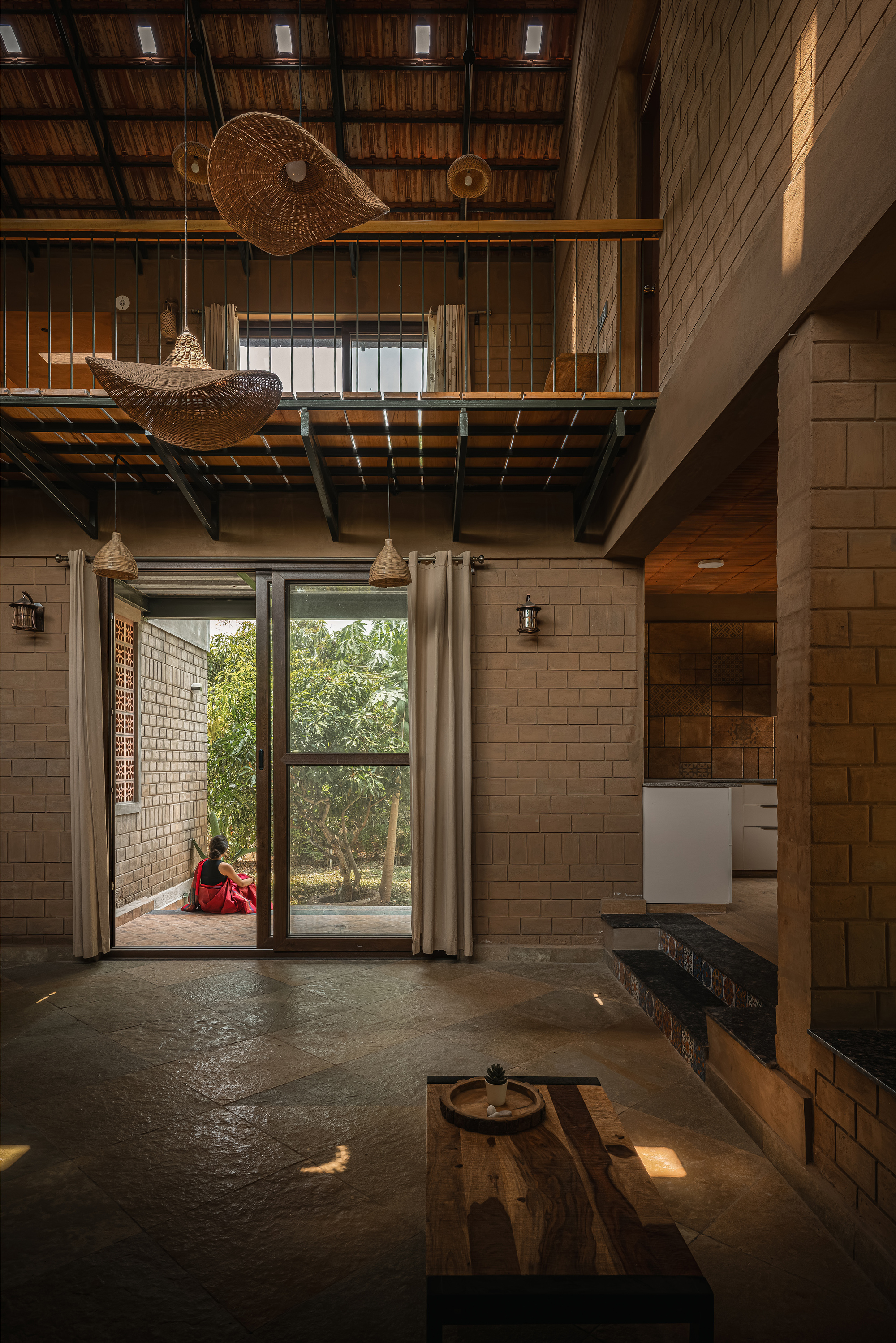 Double-height living space with a steel and timber mezzanine, woven pendant lamps and a glazed door opening to a shaded garden