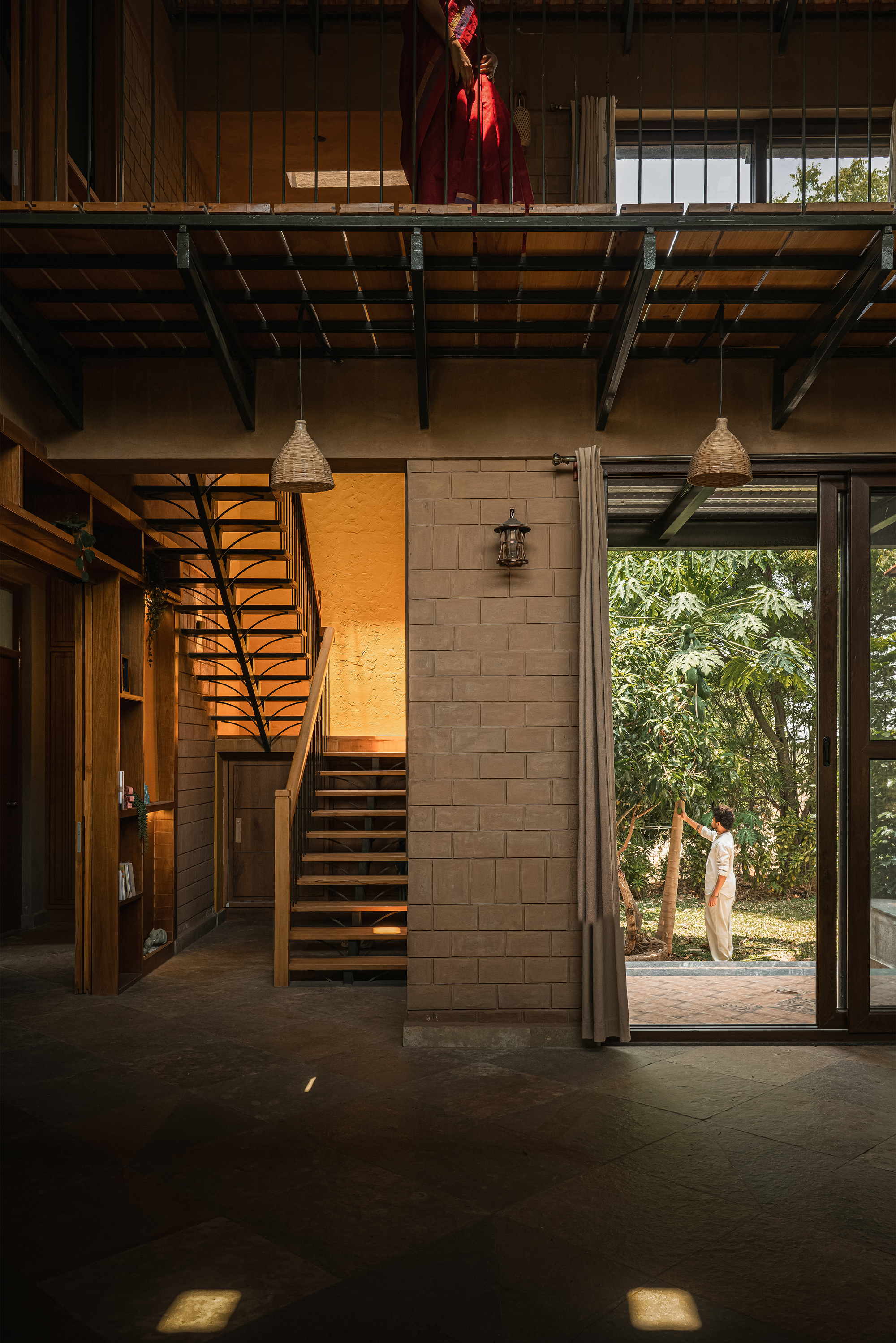 Double-height living space with open timber stair, brick pier and wicker pendants, a person standing at the open glass door to the garden