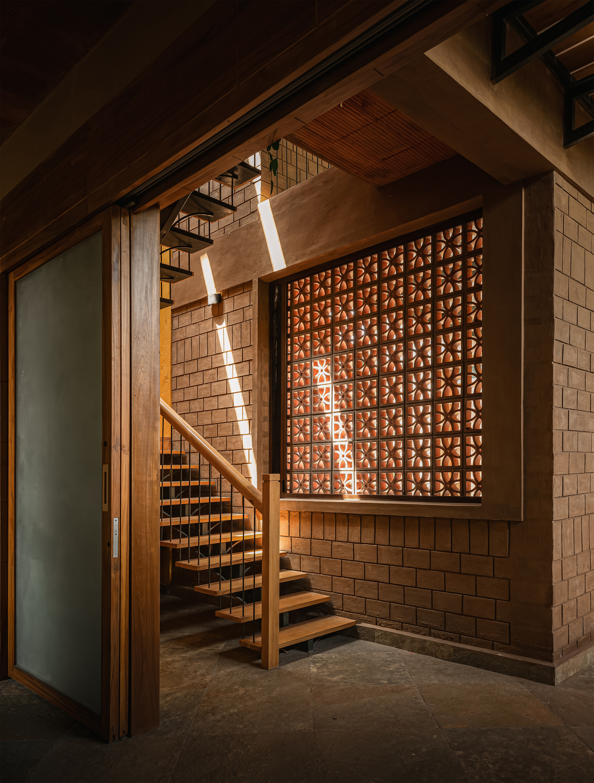 Timber stair beside a floral terracotta jali screen, with a narrow shaft of sunlight crossing the brick wall