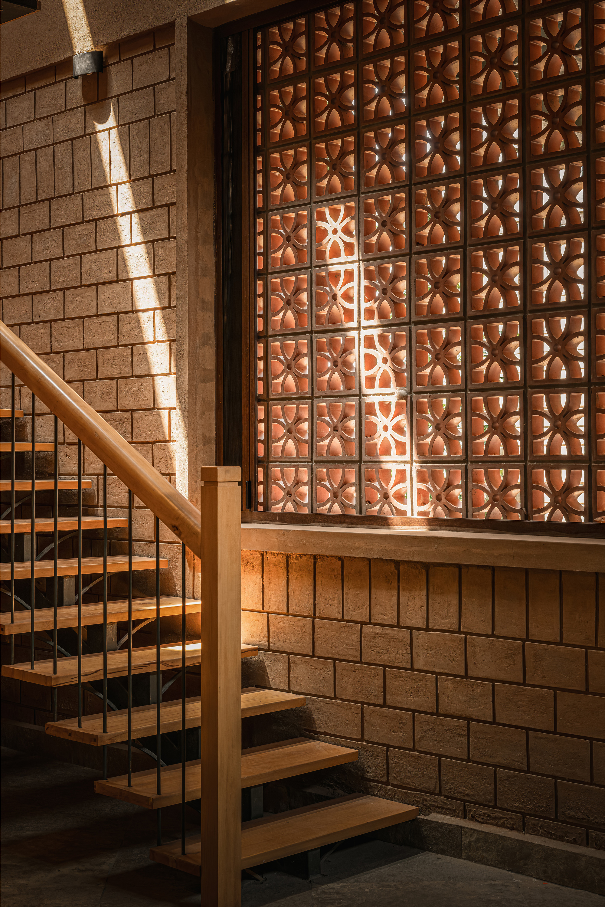Close-up of the terracotta jali screen above a timber handrail, with a diagonal blade of afternoon light on the brick