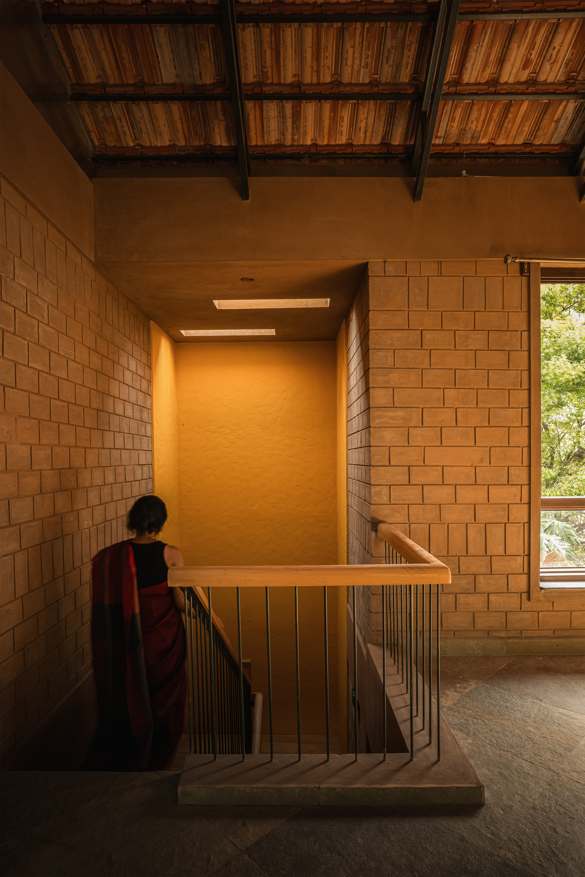 Stair landing with a yellow plastered wall and timber rail, a woman in a red sari descending beside brick walls under a tiled roof