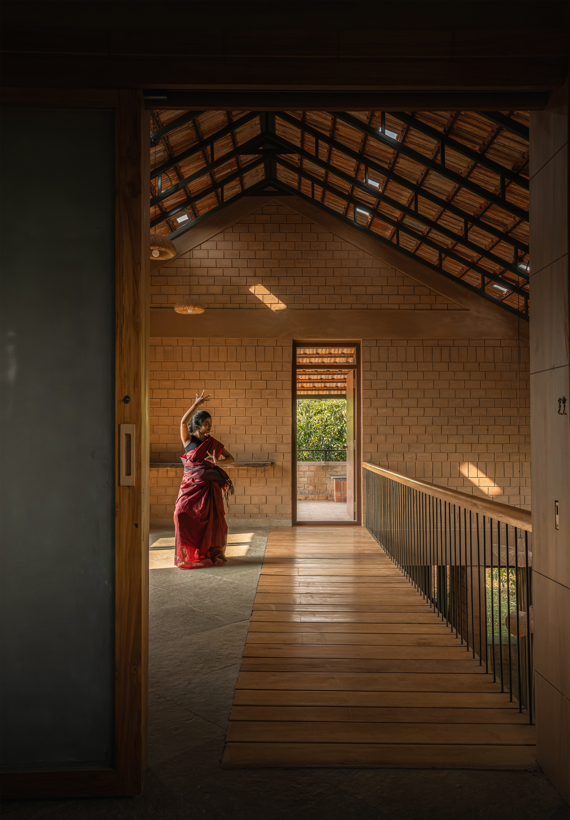 Timber-floored bridge leading to a brick gabled room with exposed steel roof trusses, a dancer in a red sari lit by patches of sun