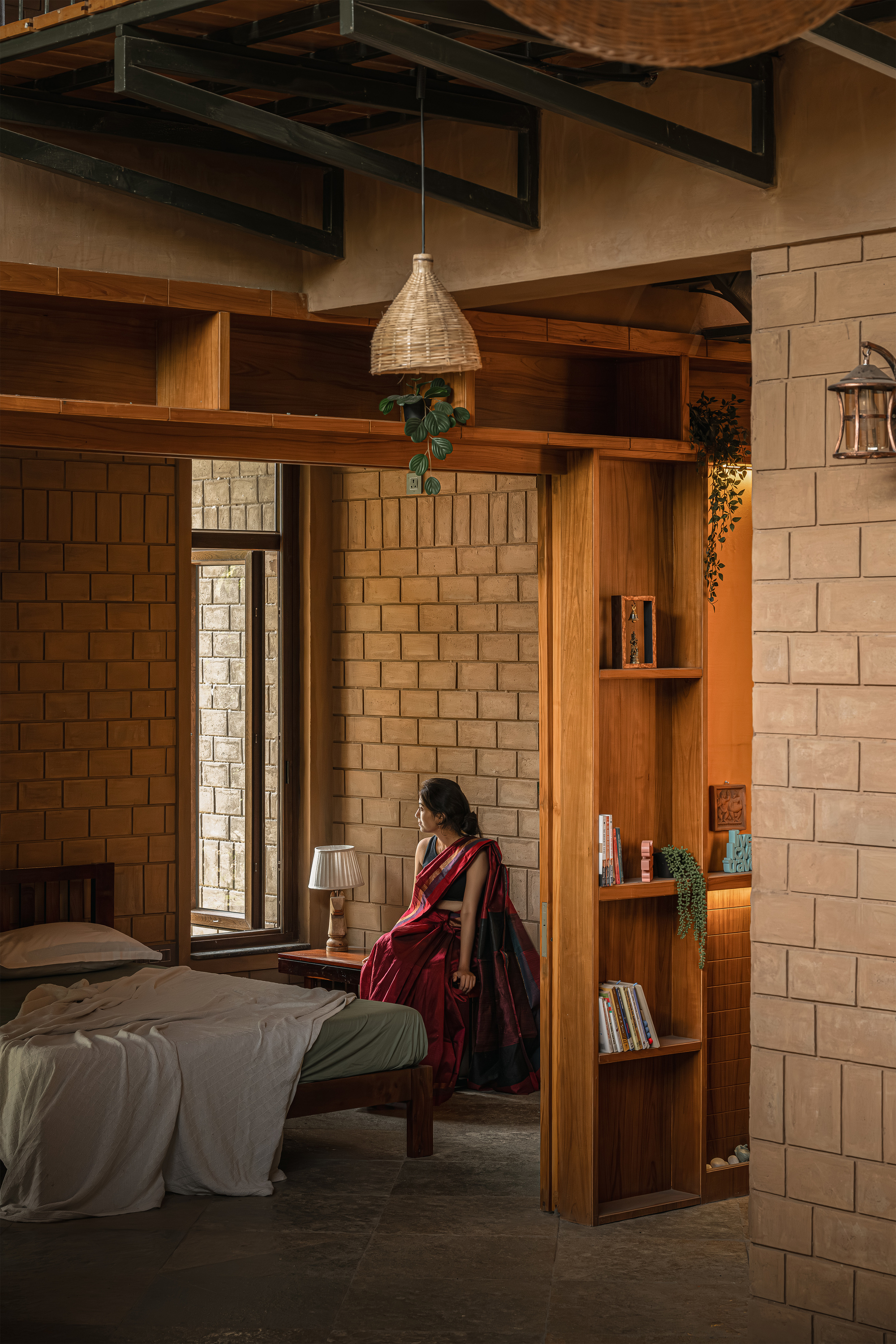 Woman in a red sari seated beside a built-in timber bookshelf, with a woven pendant lamp hanging over the bed