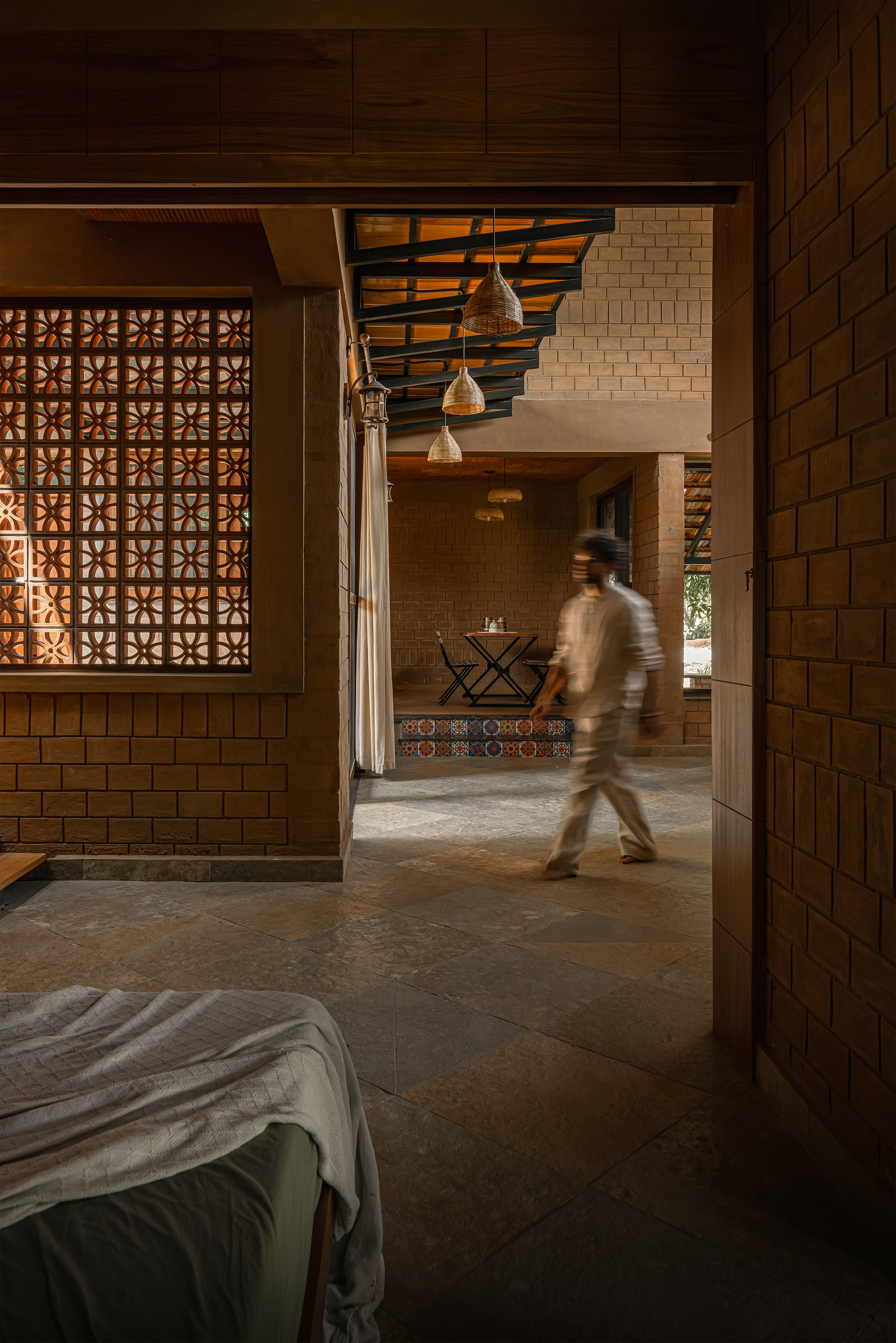 Blurred figure walking past a patterned jali screen toward a dining table, with woven pendant lamps hanging below steel roof framing
