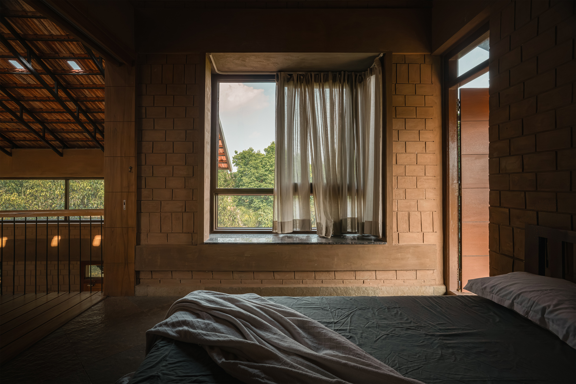 Bedroom window framed in exposed brick with sheer curtains, looking onto green treetops, with a tiled roof visible at left