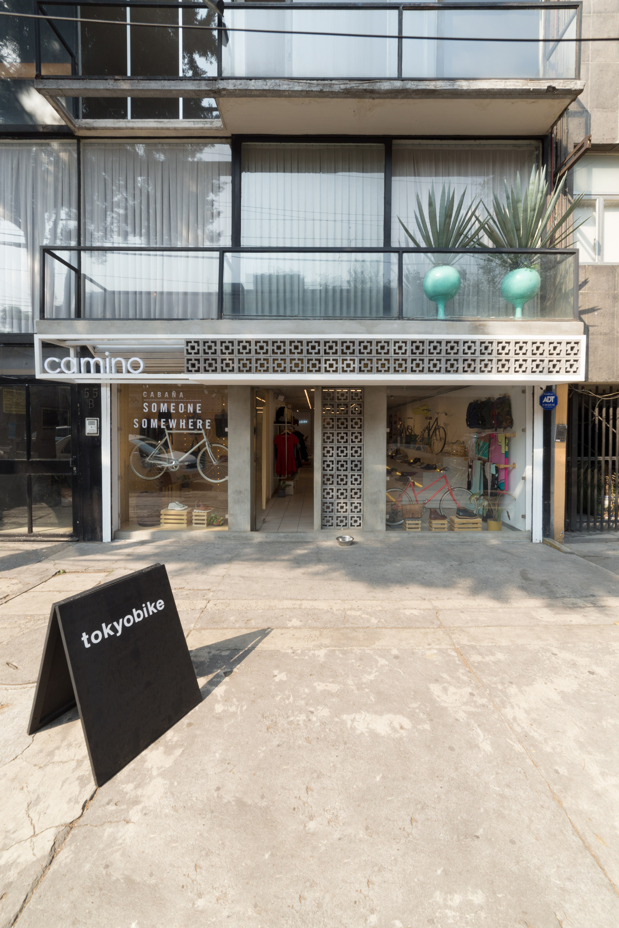 Storefront with a perforated concrete block screen above the entrance, planted pots on the balcony and an A-frame sign on sunlit pavement