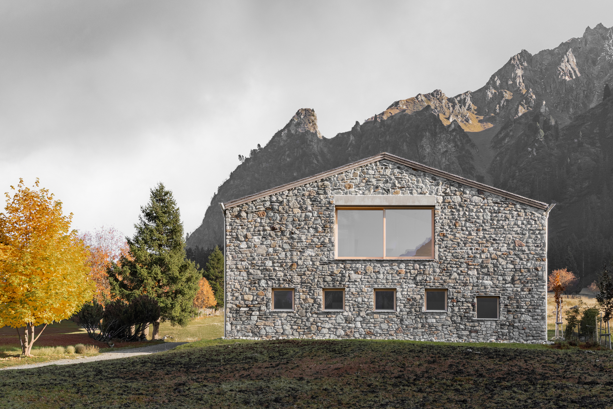 Gabled fieldstone facade with a large timber-framed window and small square openings, set against a rocky peak under an overcast autumn sky