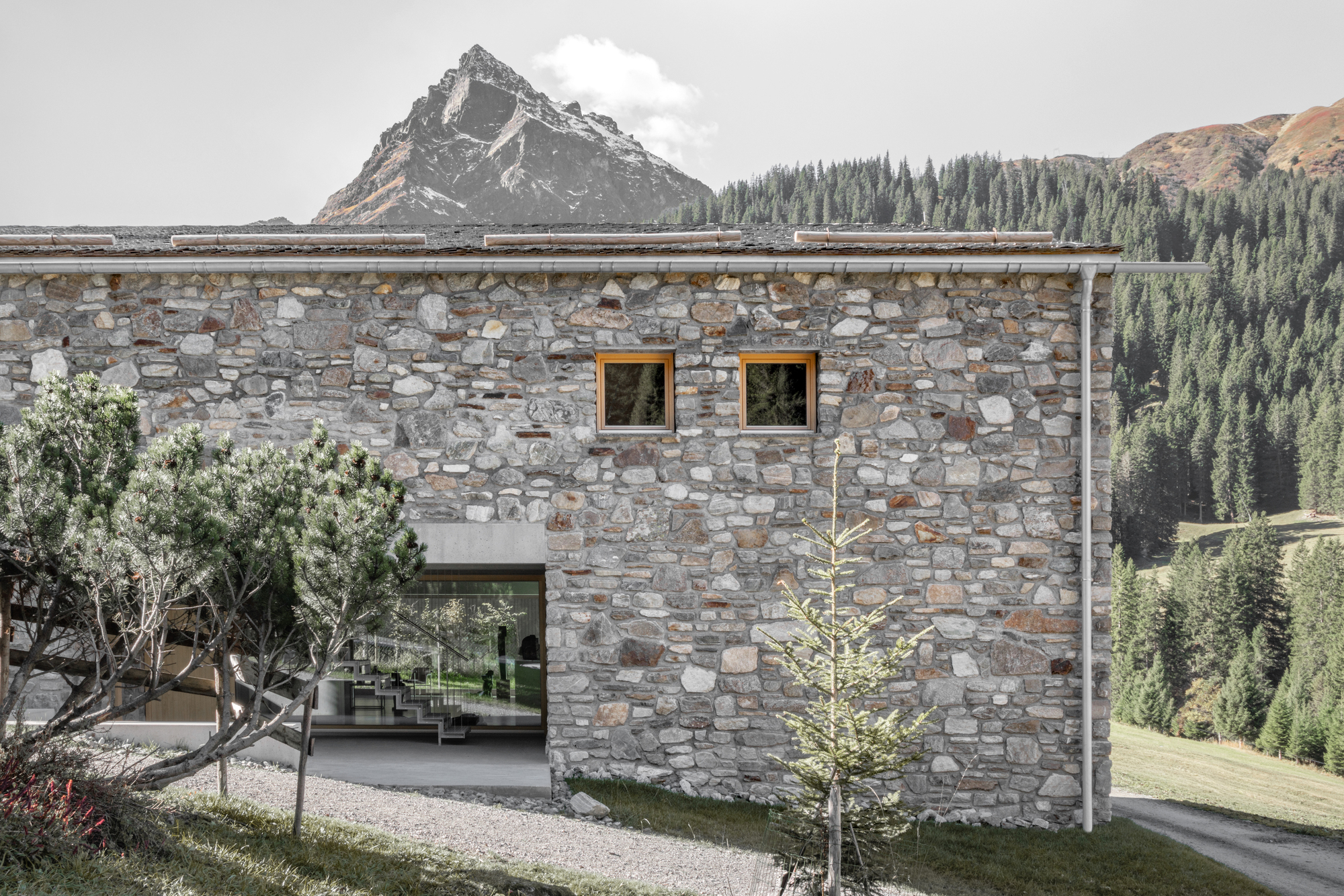 Side elevation of the rubble stone wall with two small timber-framed windows and an open glazed corner showing a staircase, with a pointed peak behind