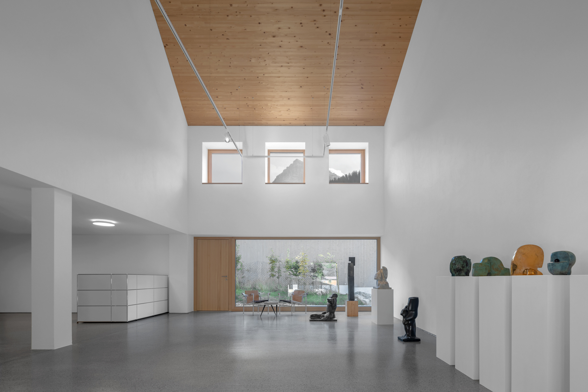 Double-height white gallery with a timber ceiling, clerestory windows and sculptures on plinths, lit by daylight from a large glazed opening