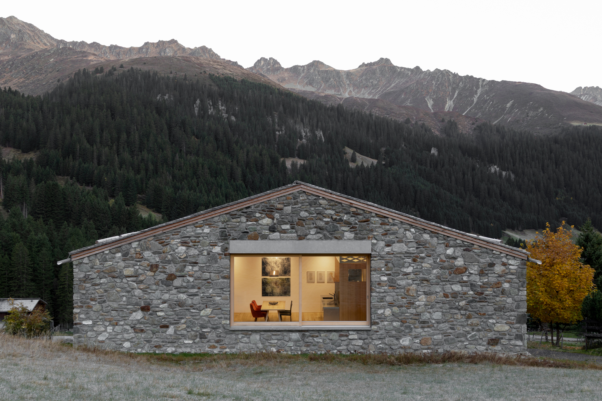 Stone gable end with a large lit window revealing a dining room, glowing at dusk against a forested mountain slope and frosted grass