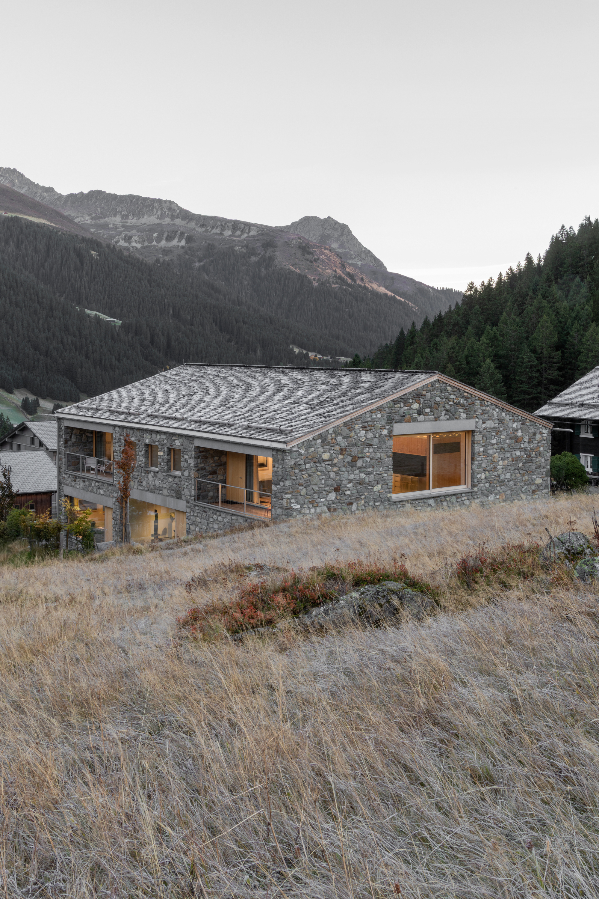 Stone-clad house with a slate roof and a large corner window on a grassy slope at dusk