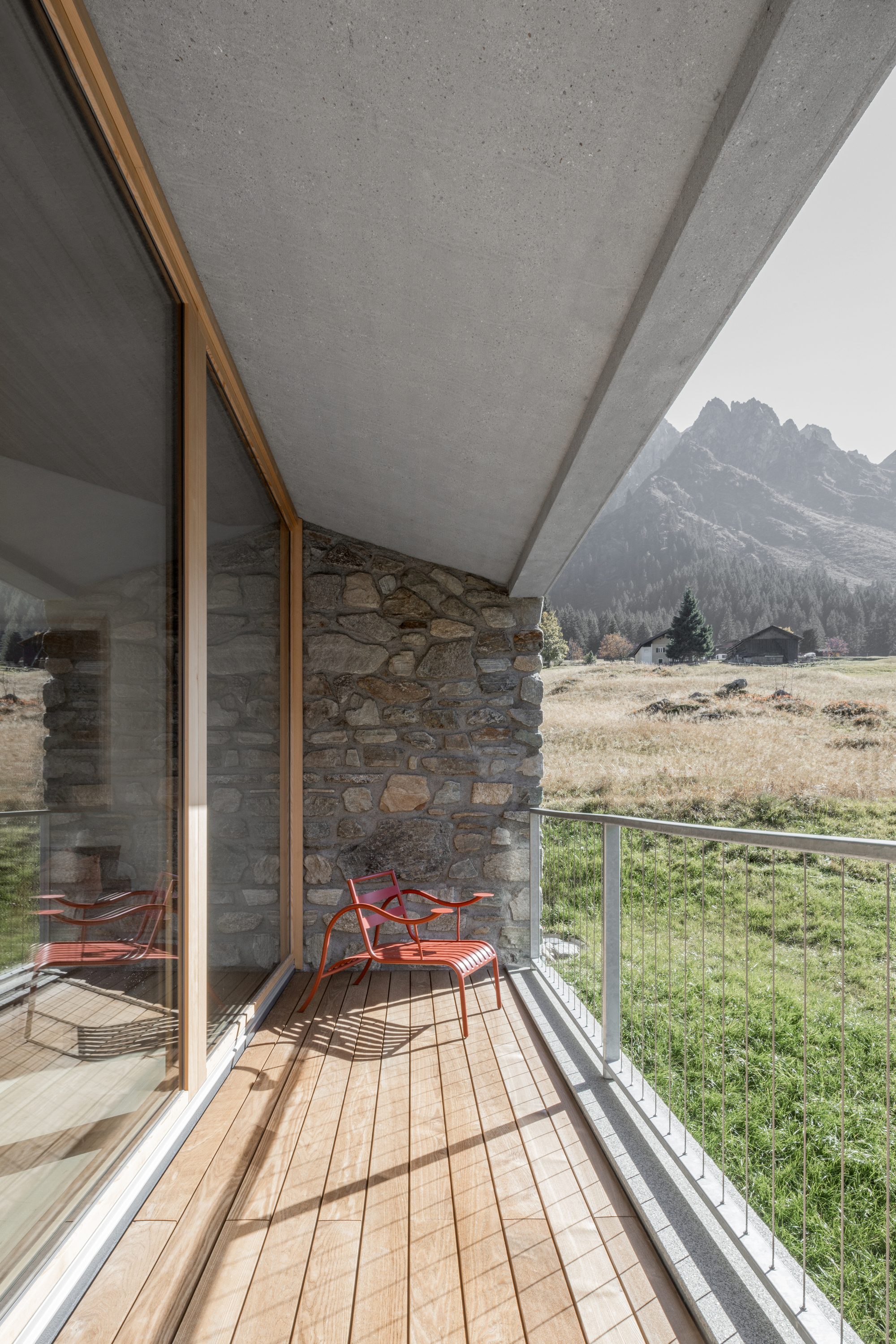 Timber-floored balcony with a red chair, stone wall and concrete soffit facing a sunlit alpine meadow