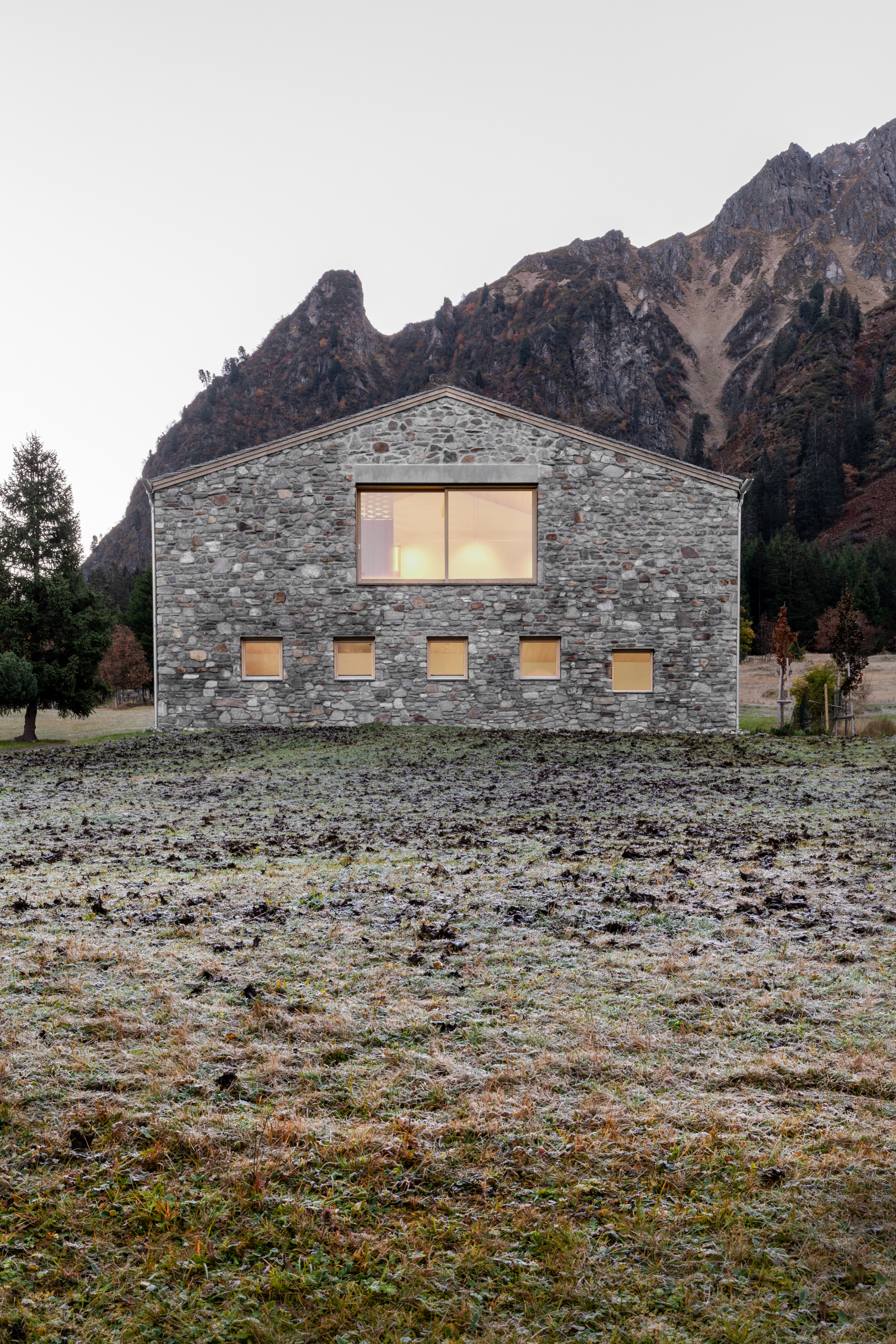 Gabled stone end wall with a large lit window and five small square openings above frosted grass