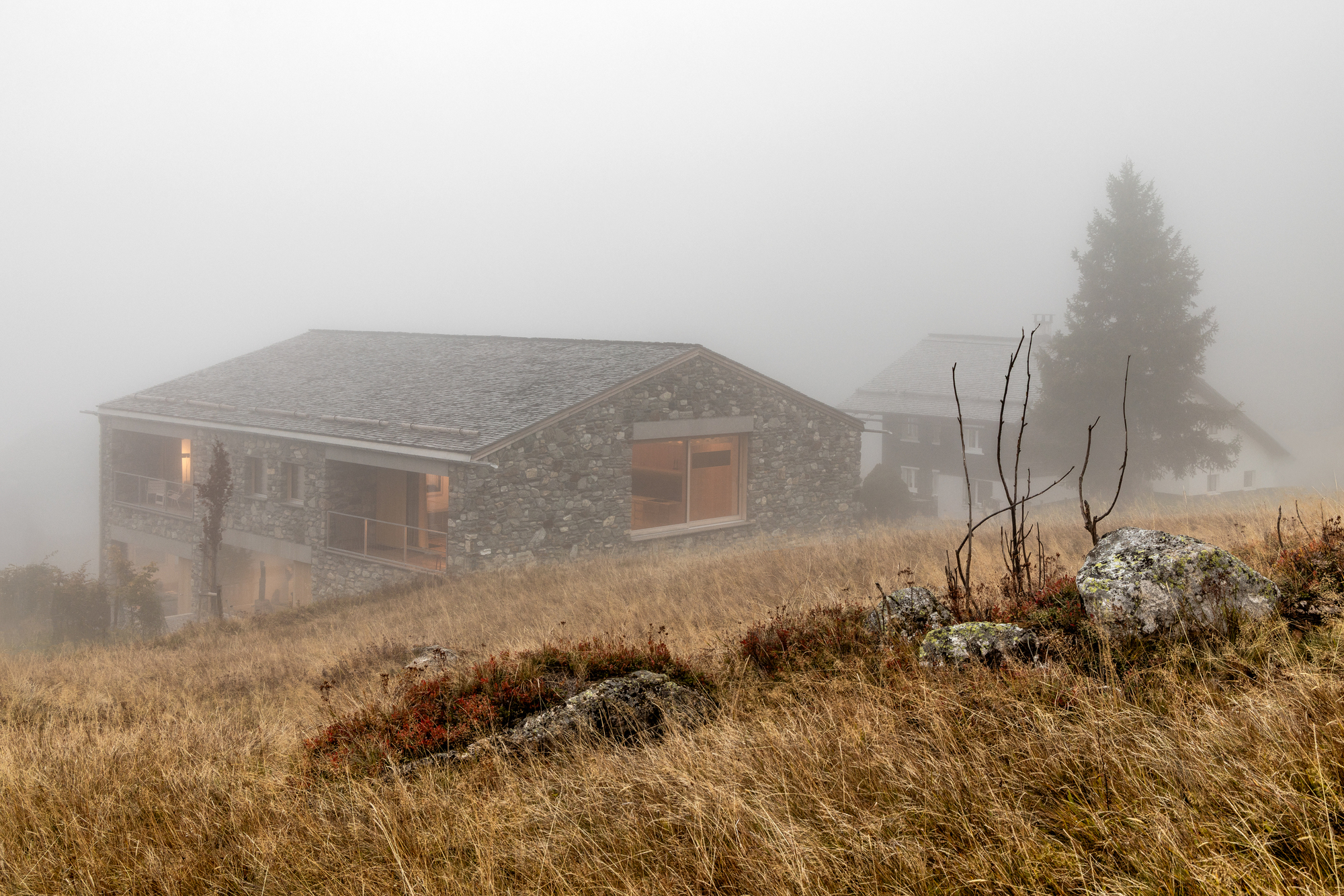Stone house with a slate roof half hidden in thick fog above rust-colored grass and boulders
