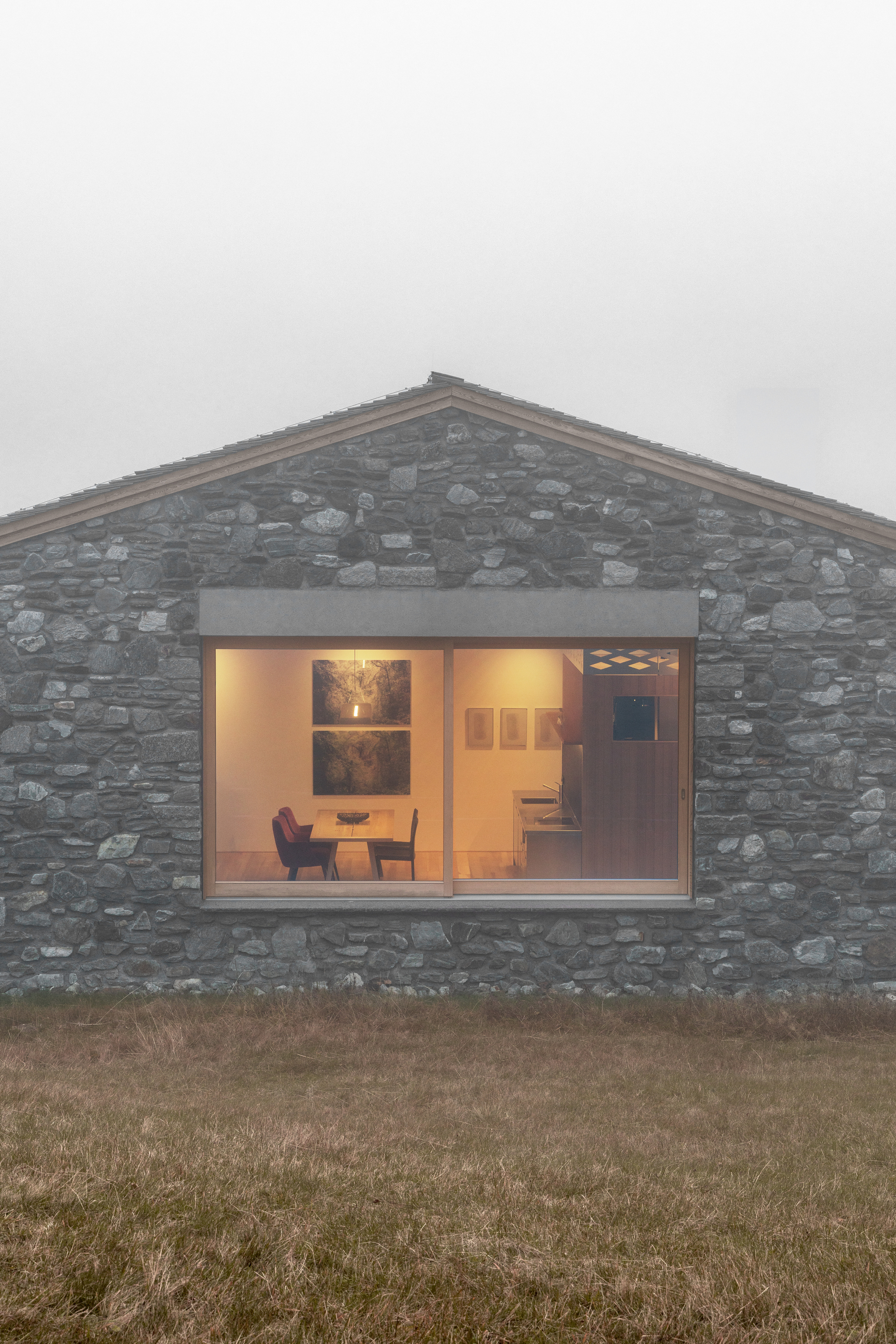Rubble stone gable end with a lit timber-framed picture window showing a dining table, seen through low fog over grass