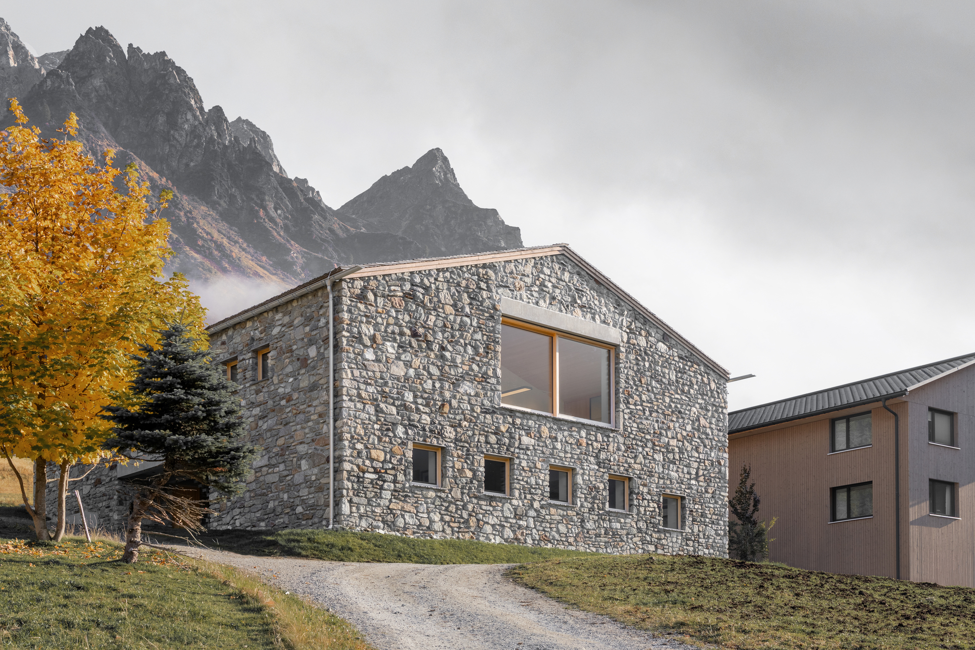 Stone gabled house on a gravel track with small timber windows, an oversized upper window and a yellow larch before misty peaks