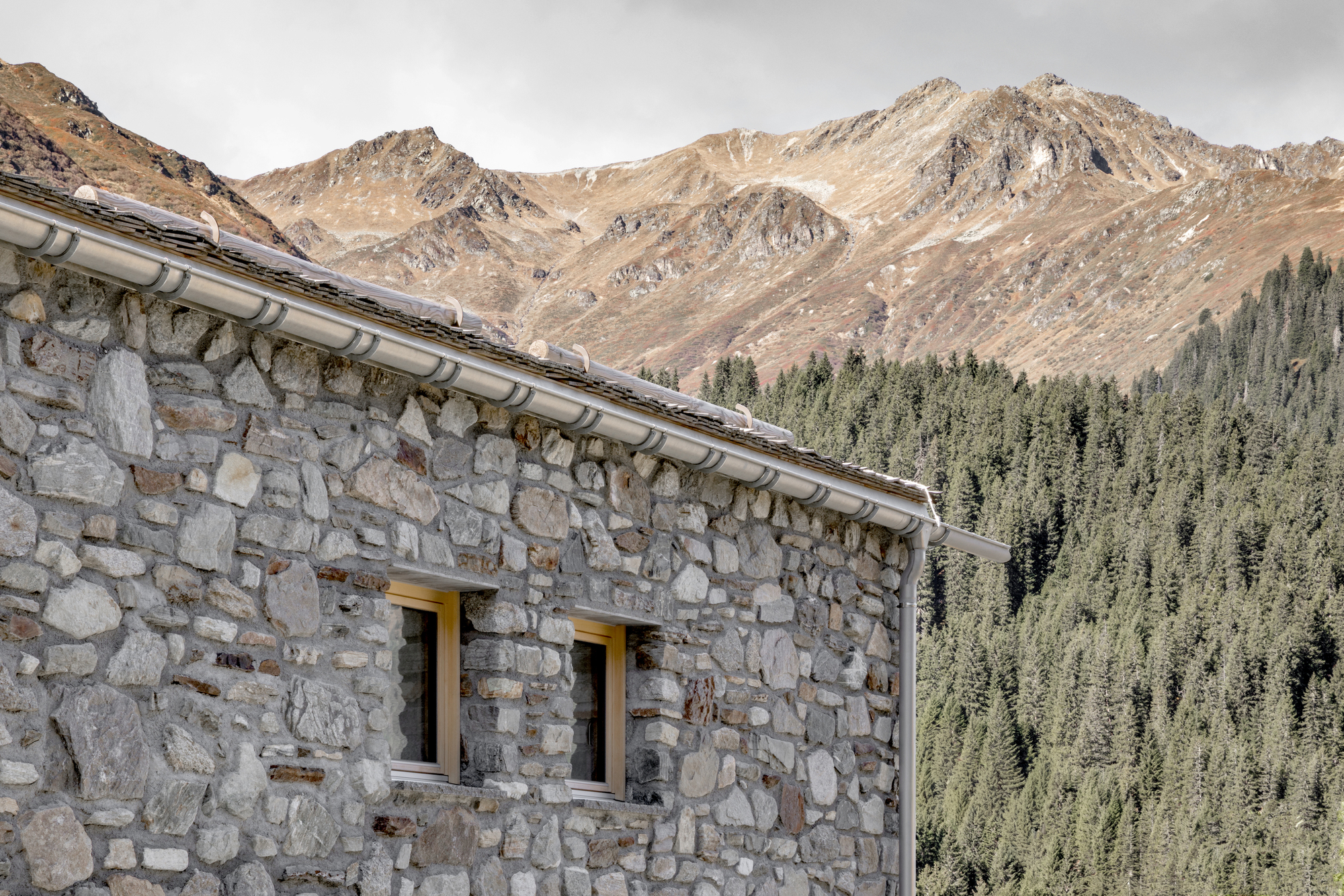 Corner of the stone wall with two small timber-framed windows and a metal gutter, set against brown mountain slopes and spruce forest