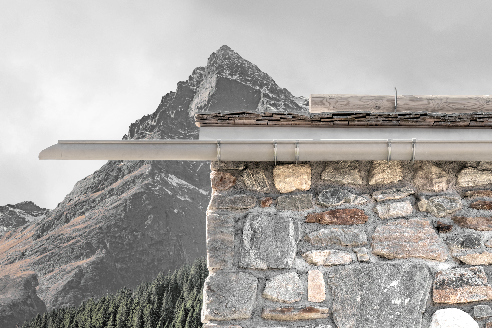 Close-up of the metal gutter and slate eaves above a rough stone wall, with a rocky peak rising in overcast light