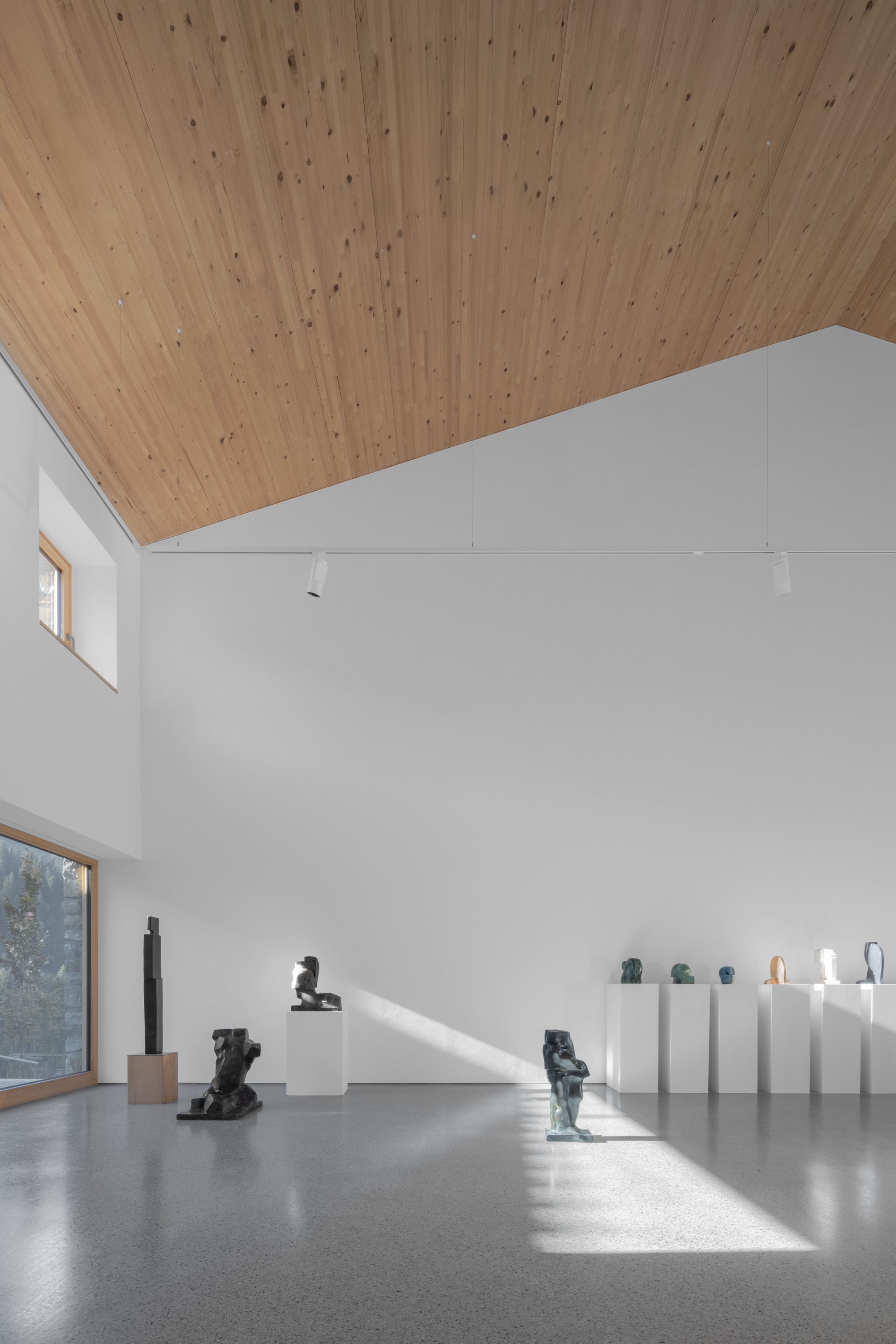 Double-height gallery with sculptures on white plinths, a sloped timber ceiling and a diagonal band of sunlight across the concrete floor