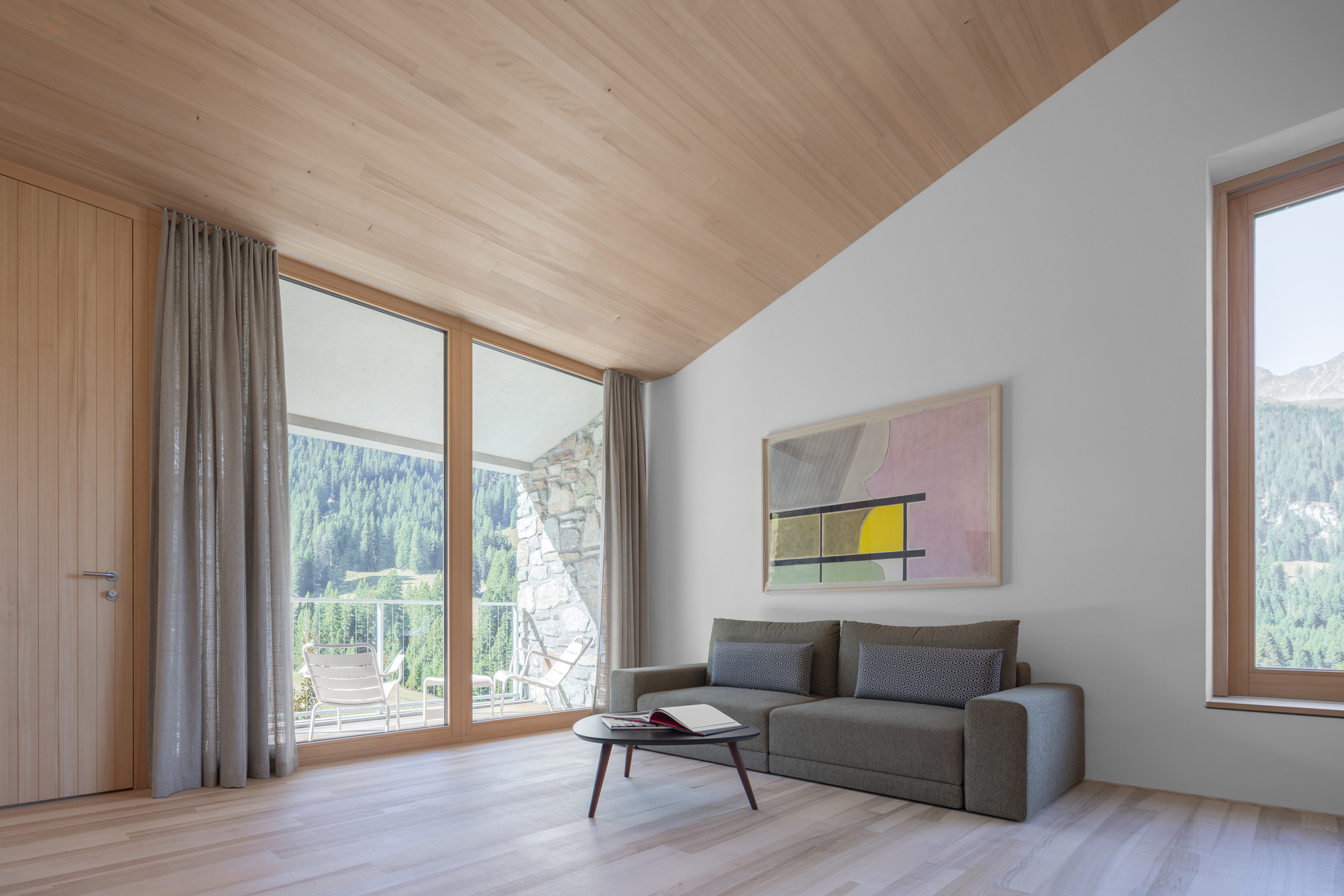 Living room with grey sofa under a sloped timber ceiling, full-height window opening onto a balcony and forested slope