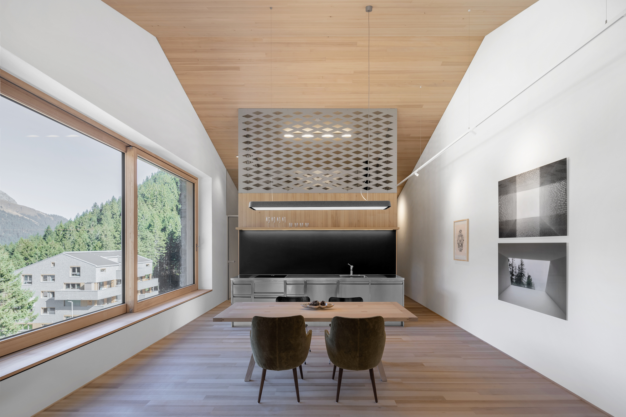 Dining table and steel kitchen block beneath a perforated metal canopy, with a large window framing pine forest and mountains