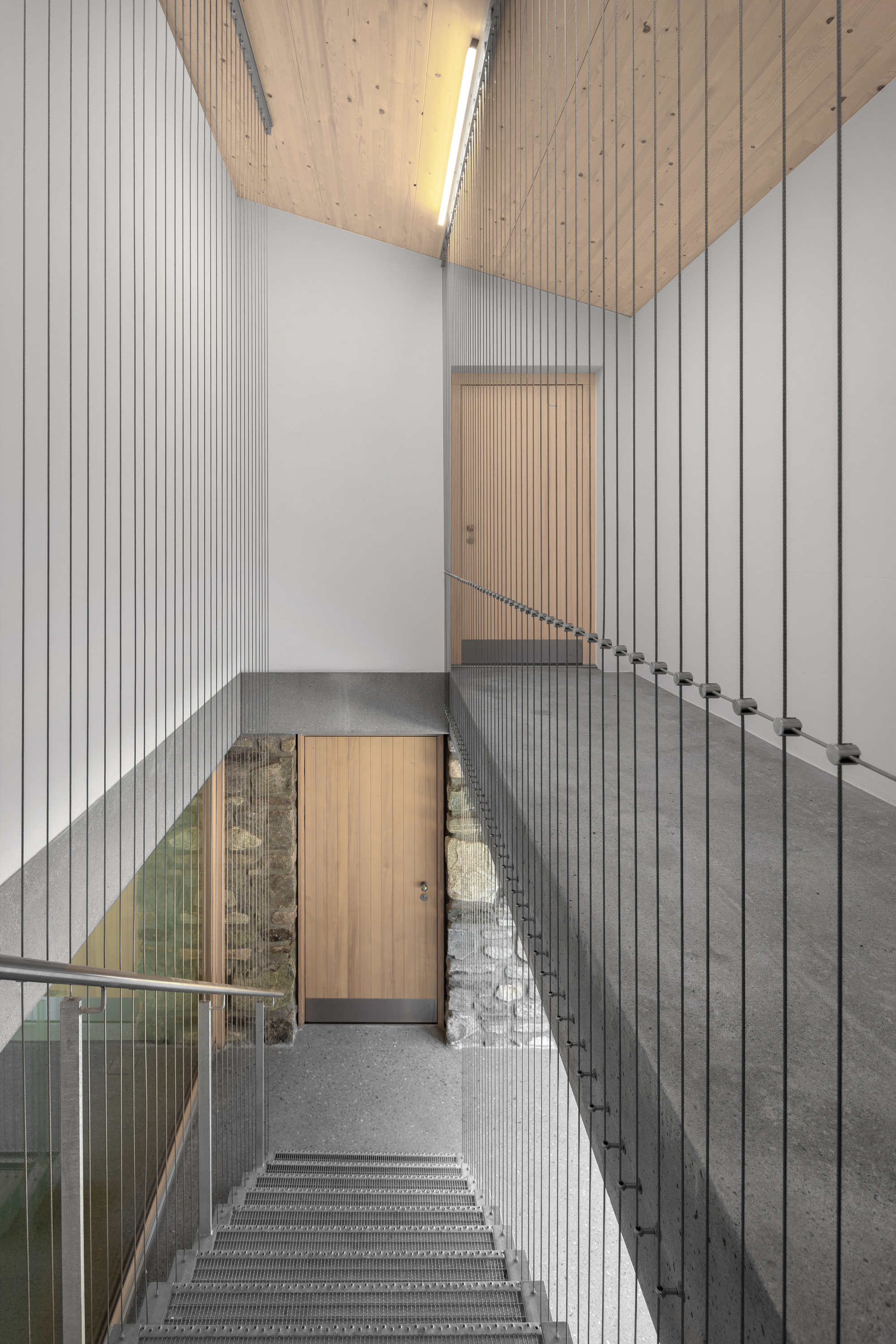 Stairwell with vertical steel cable balustrades, timber doors and an exposed rubble stone wall under a sloped wood ceiling