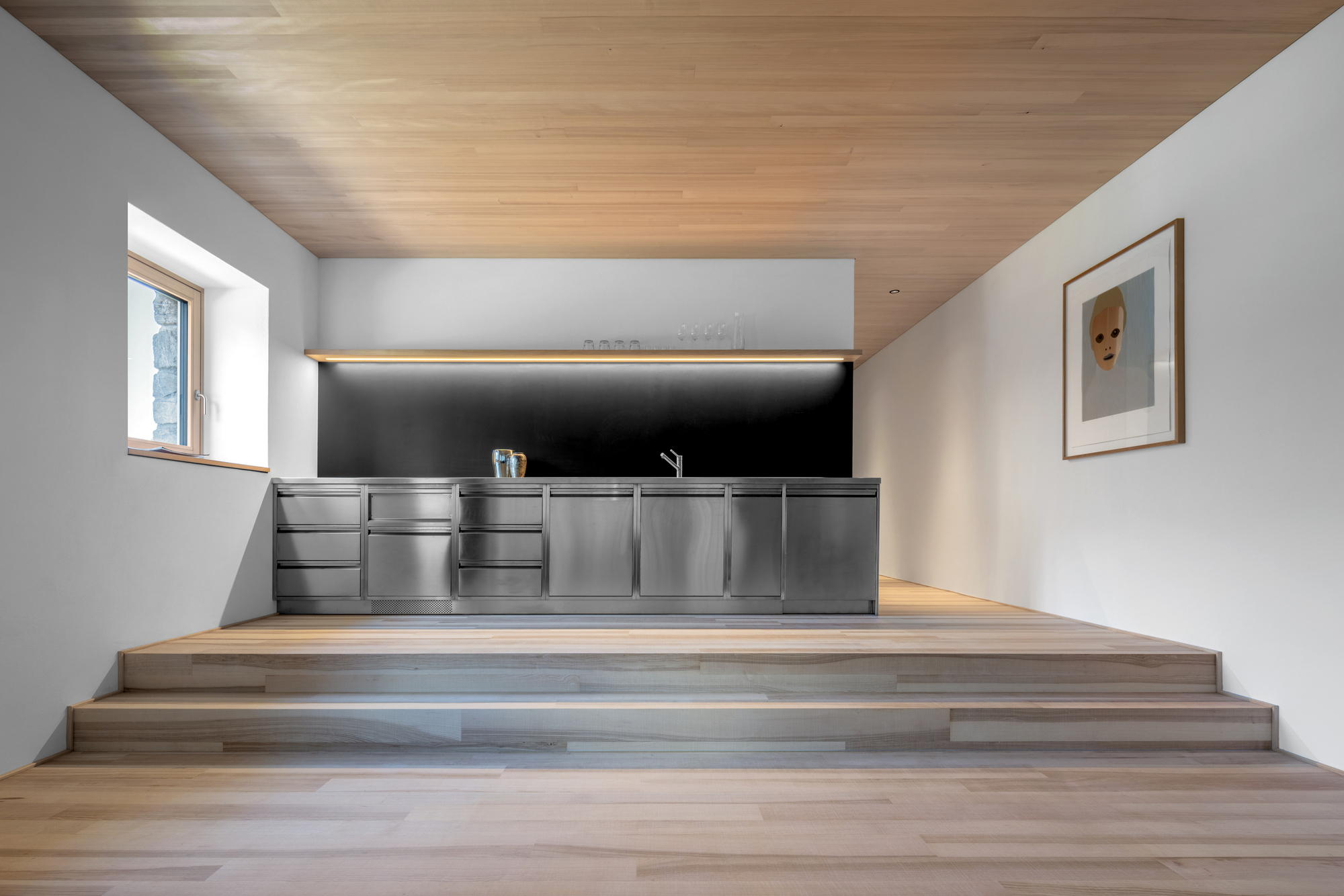 Raised kitchen platform with stainless steel cabinets, black splashback and a framed portrait on the wall in soft daylight