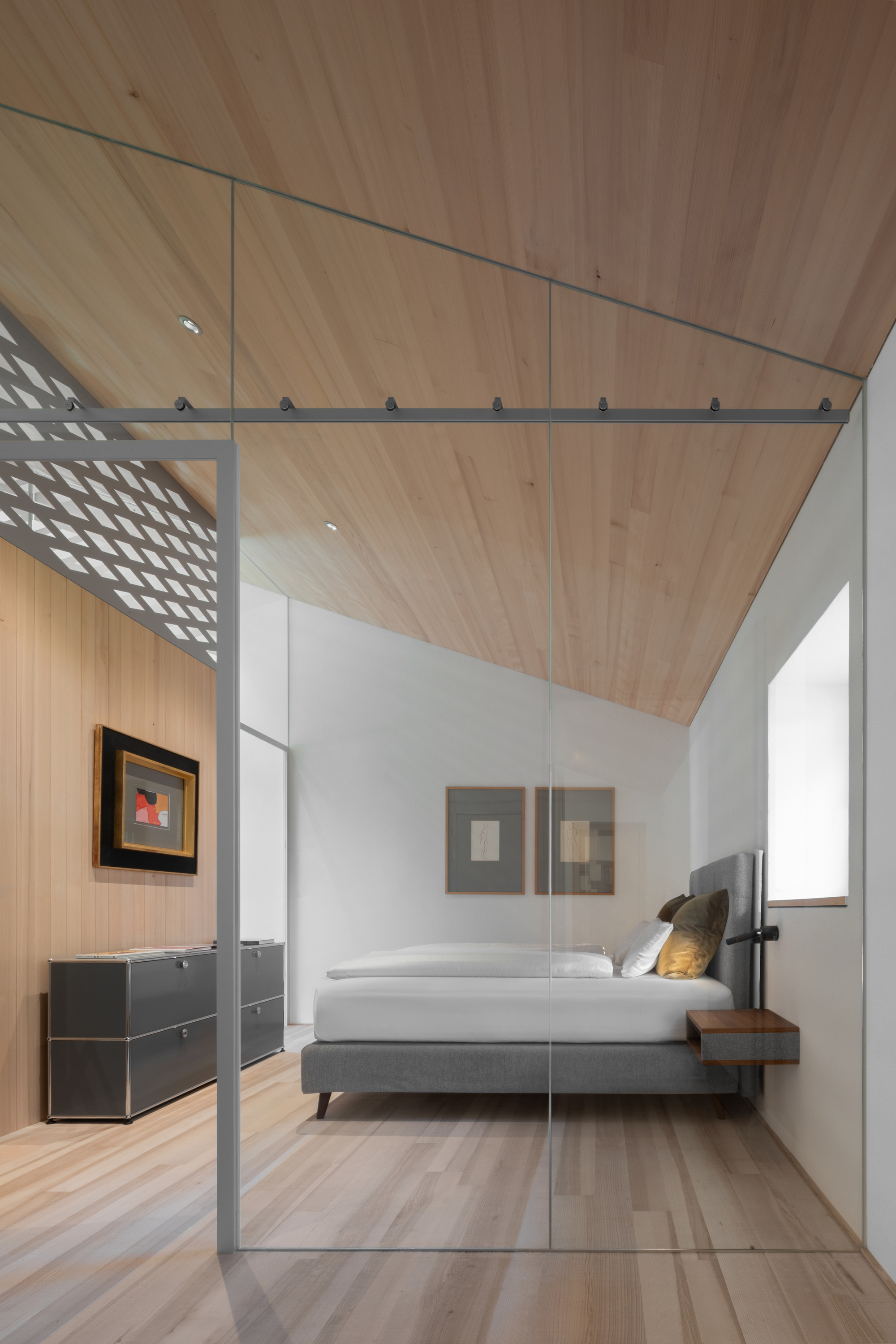 Bedroom seen through a sliding glass partition, with a sloped timber ceiling, grey upholstered bed and steel chest of drawers
