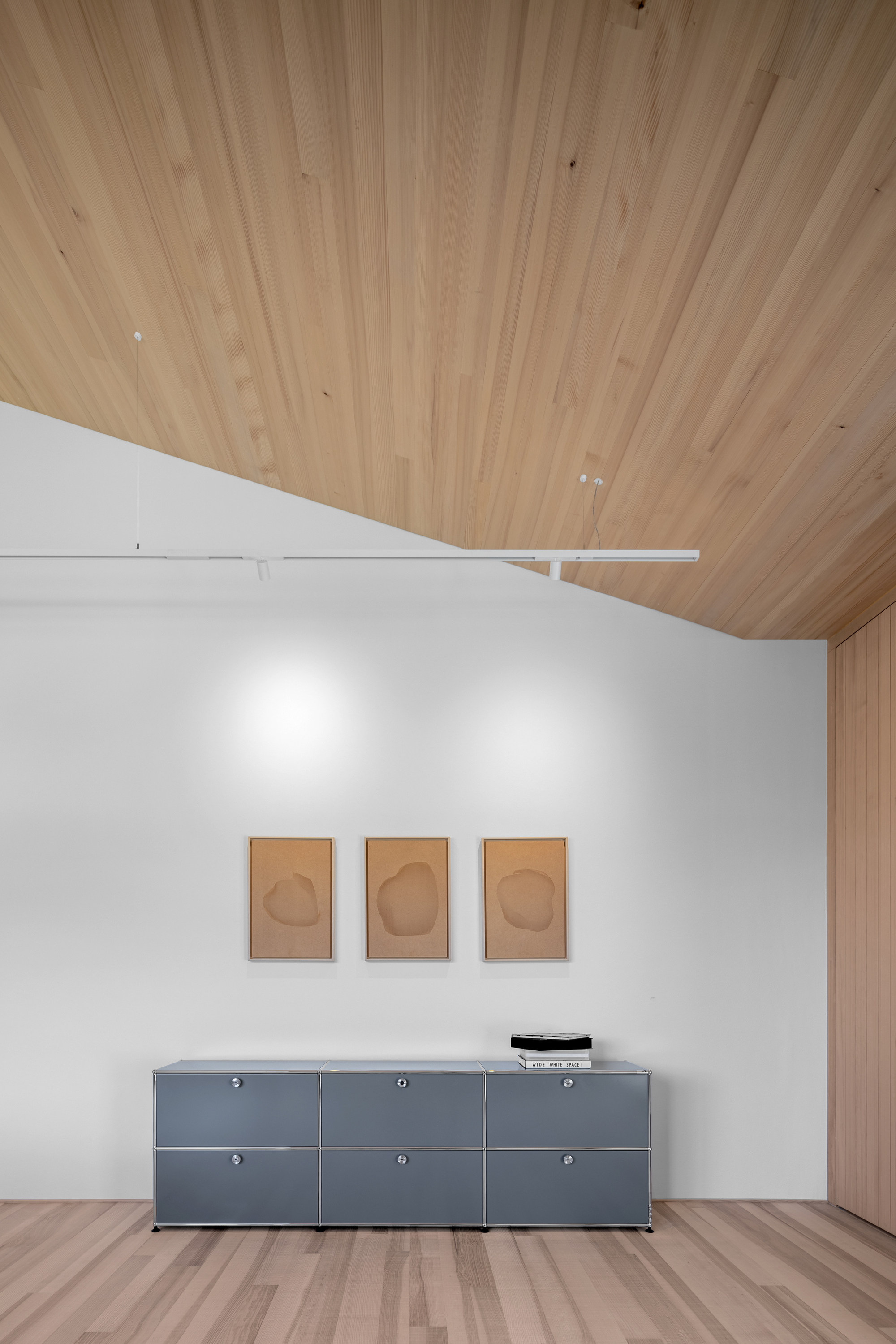 Gray steel sideboard beneath three small framed works on a white wall, with a sloped timber ceiling and a track light overhead
