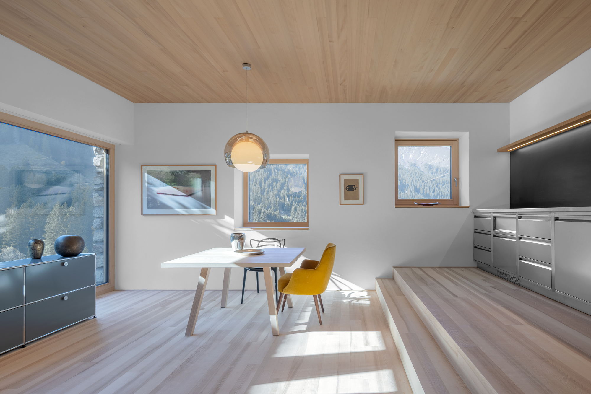 Dining area with white table and yellow chair, sunlight falling across the oak floor beside a raised kitchen platform and mountain views