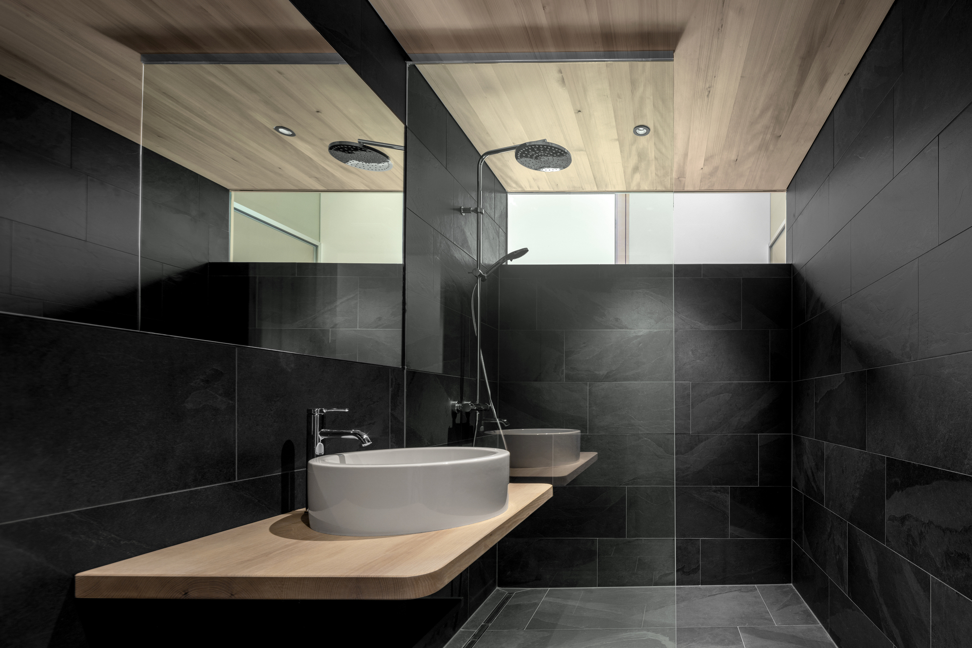 Bathroom clad in dark slate tile with a floating oak shelf, round basin and rain shower beneath a timber ceiling