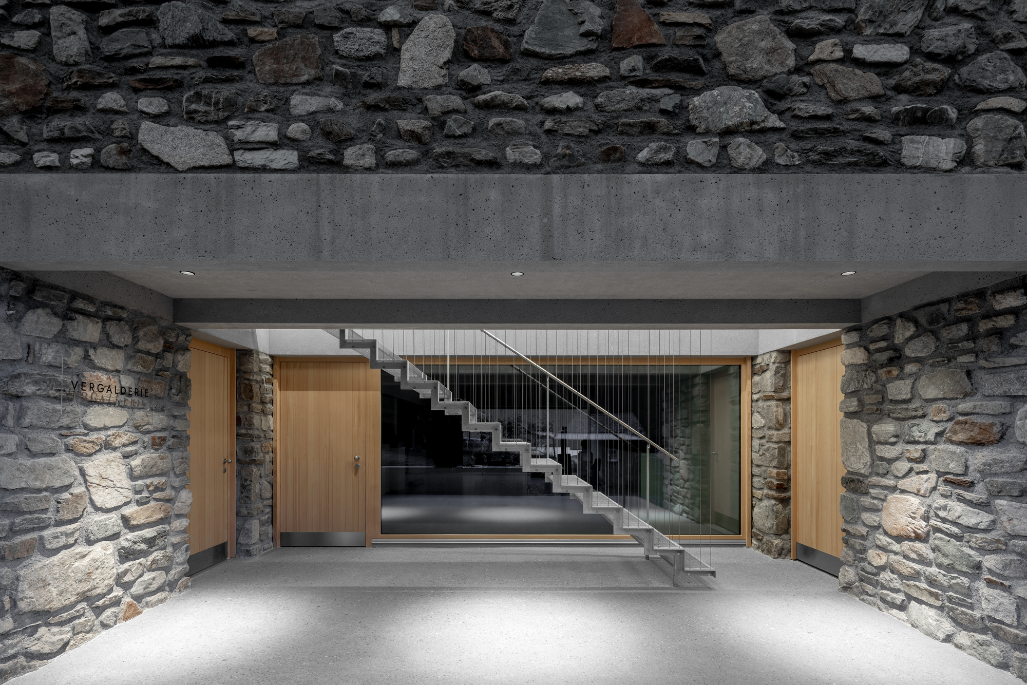 Stone-walled entrance passage with a concrete lintel, oak doors and a floating steel stair seen through the glazed wall