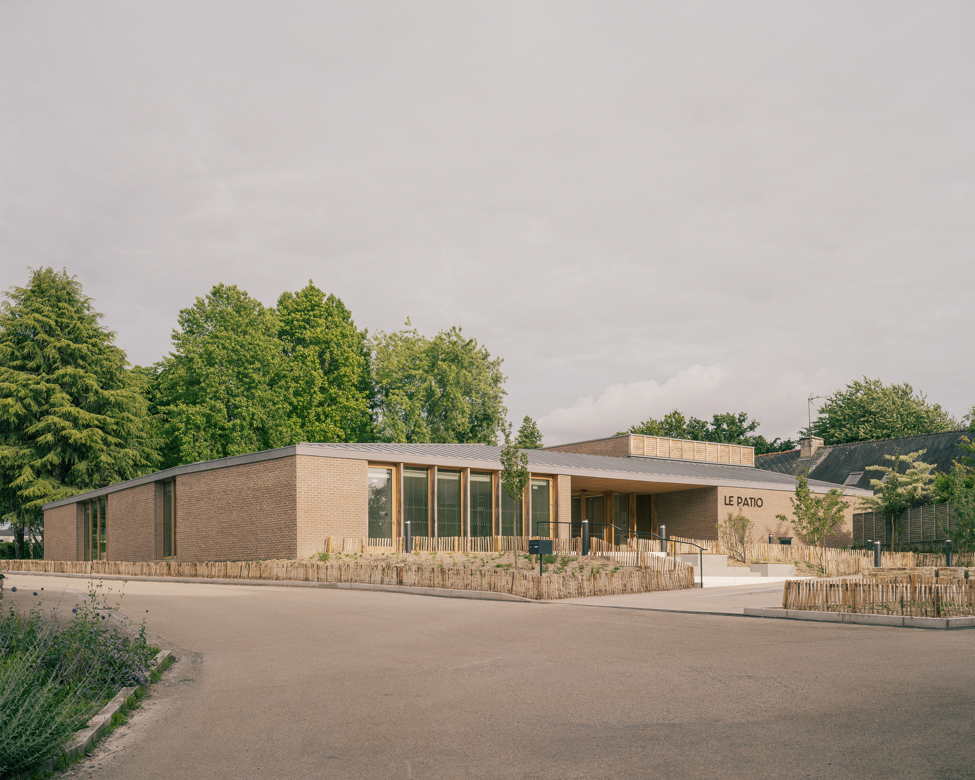 Low brick pavilion with a projecting canopy entrance and woven reed fencing along the planted edge under an overcast sky