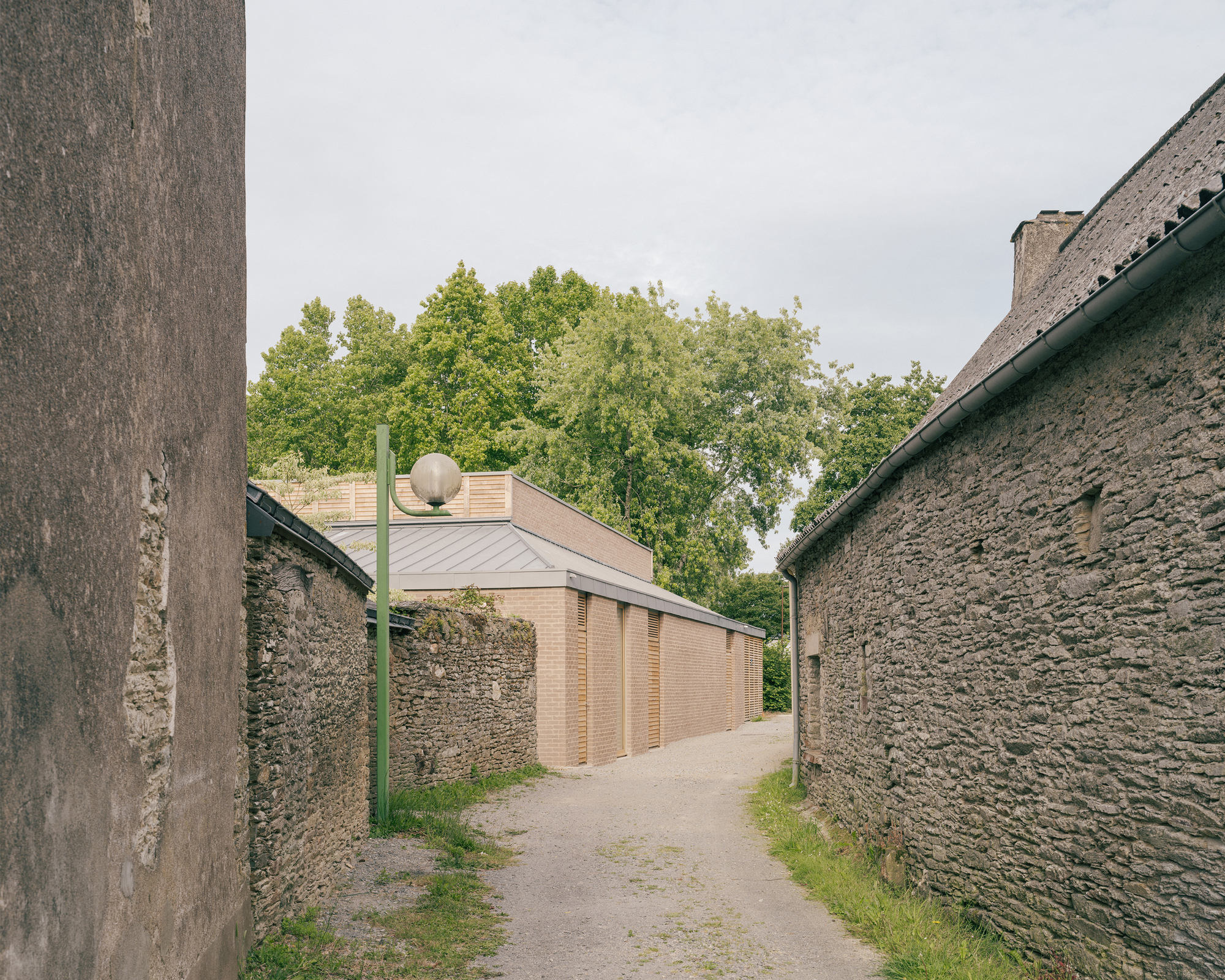 Narrow gravel lane between old stone walls, with the brick side facade and zinc roof of the pavilion beyond a green lamppost