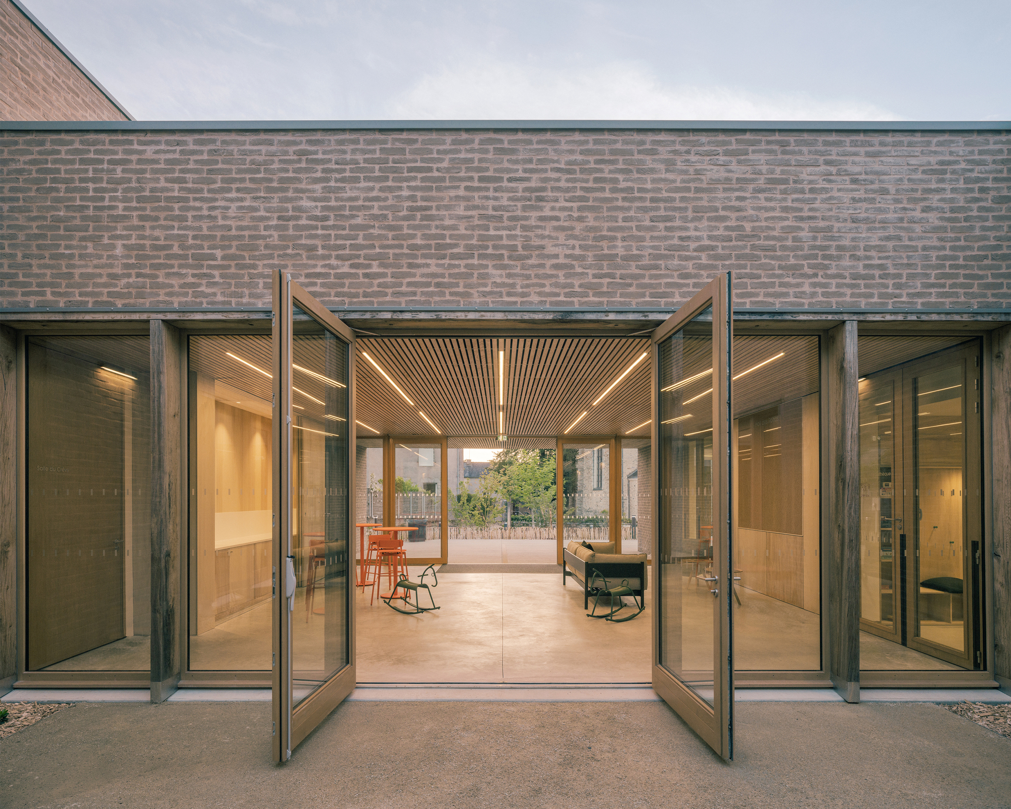 Open pivot glass doors in a timber frame beneath a brick band, revealing a slatted ceiling and furnished hall at dusk