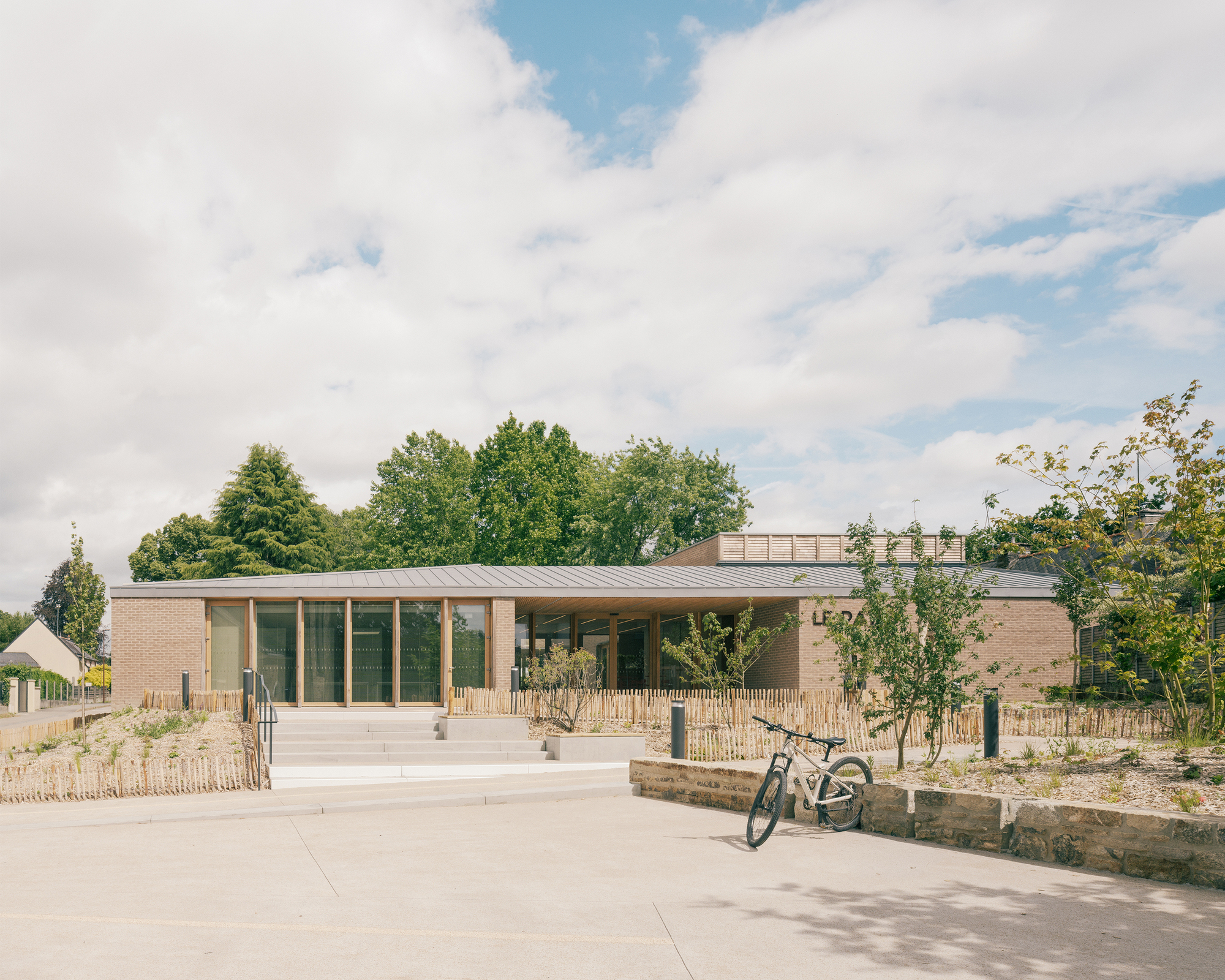 Low brick pavilion with a zinc roof and timber-framed glazing, a parked bicycle in the foreground under a cloudy summer sky