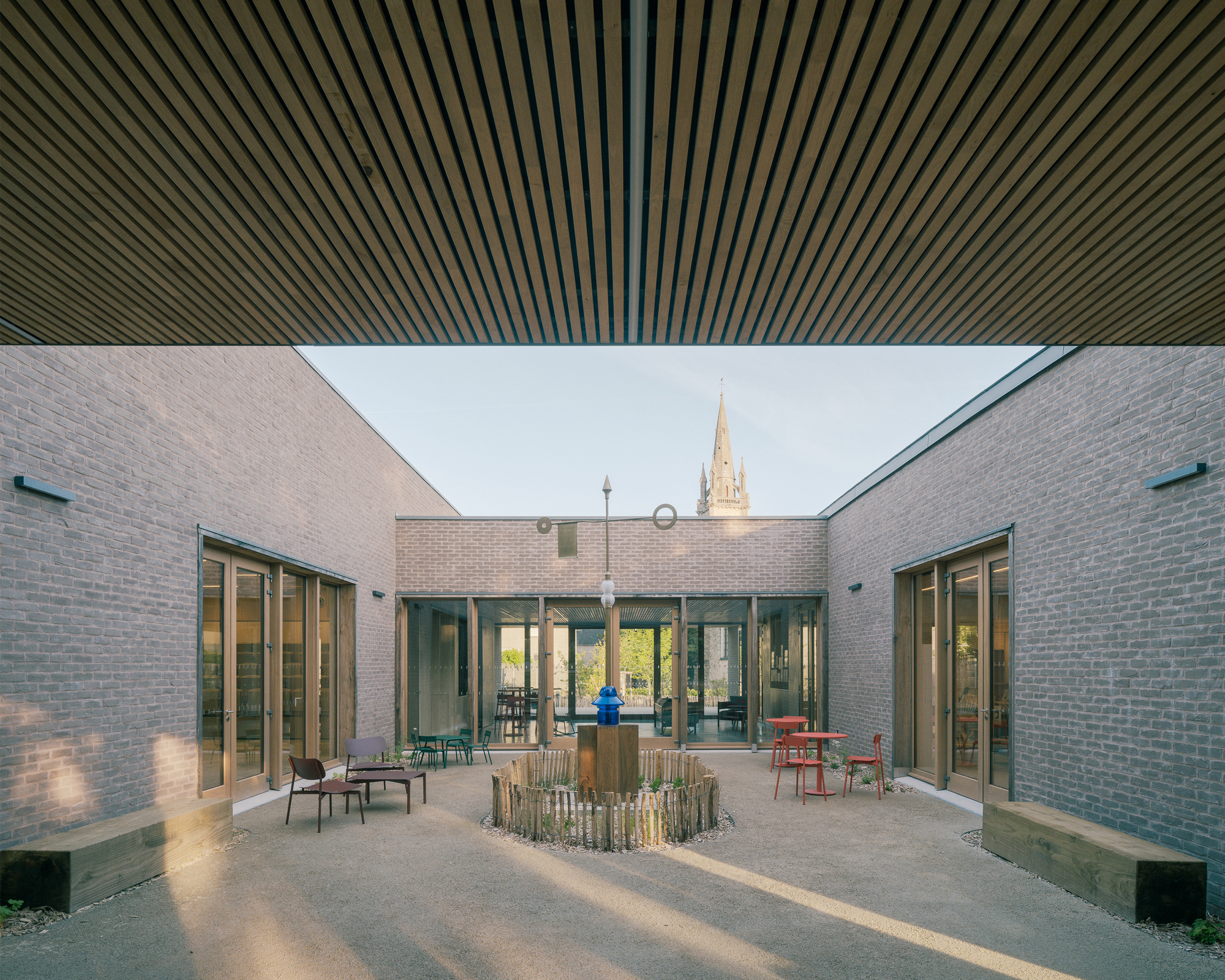 Courtyard seen from beneath the slatted timber canopy, with a church spire above the brick walls and low sun raking the gravel